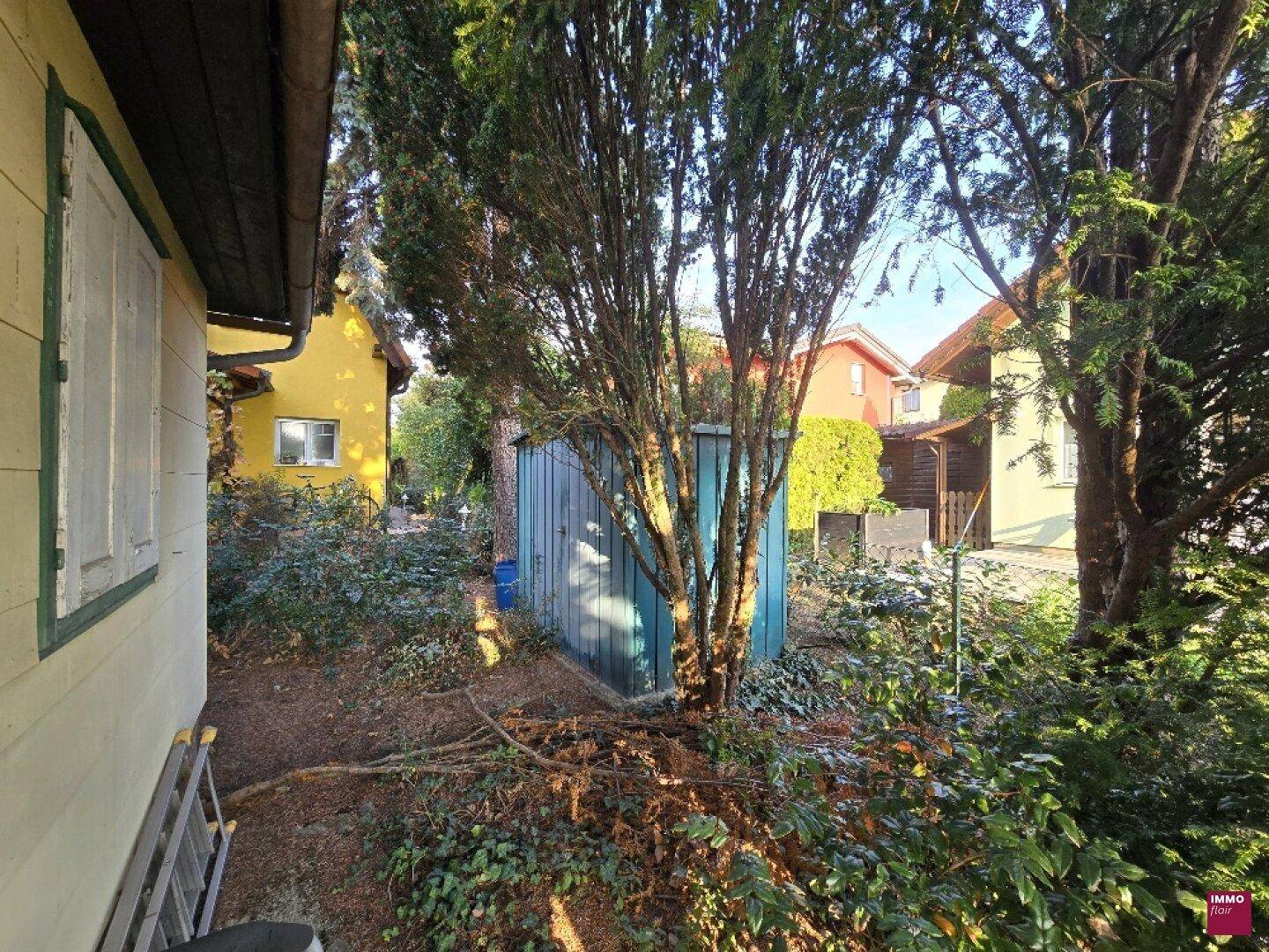 Seitenansicht des gelben Gartenhauses mit einem blauen Schuppen, umgeben von dichtem Grün und Bäumen.