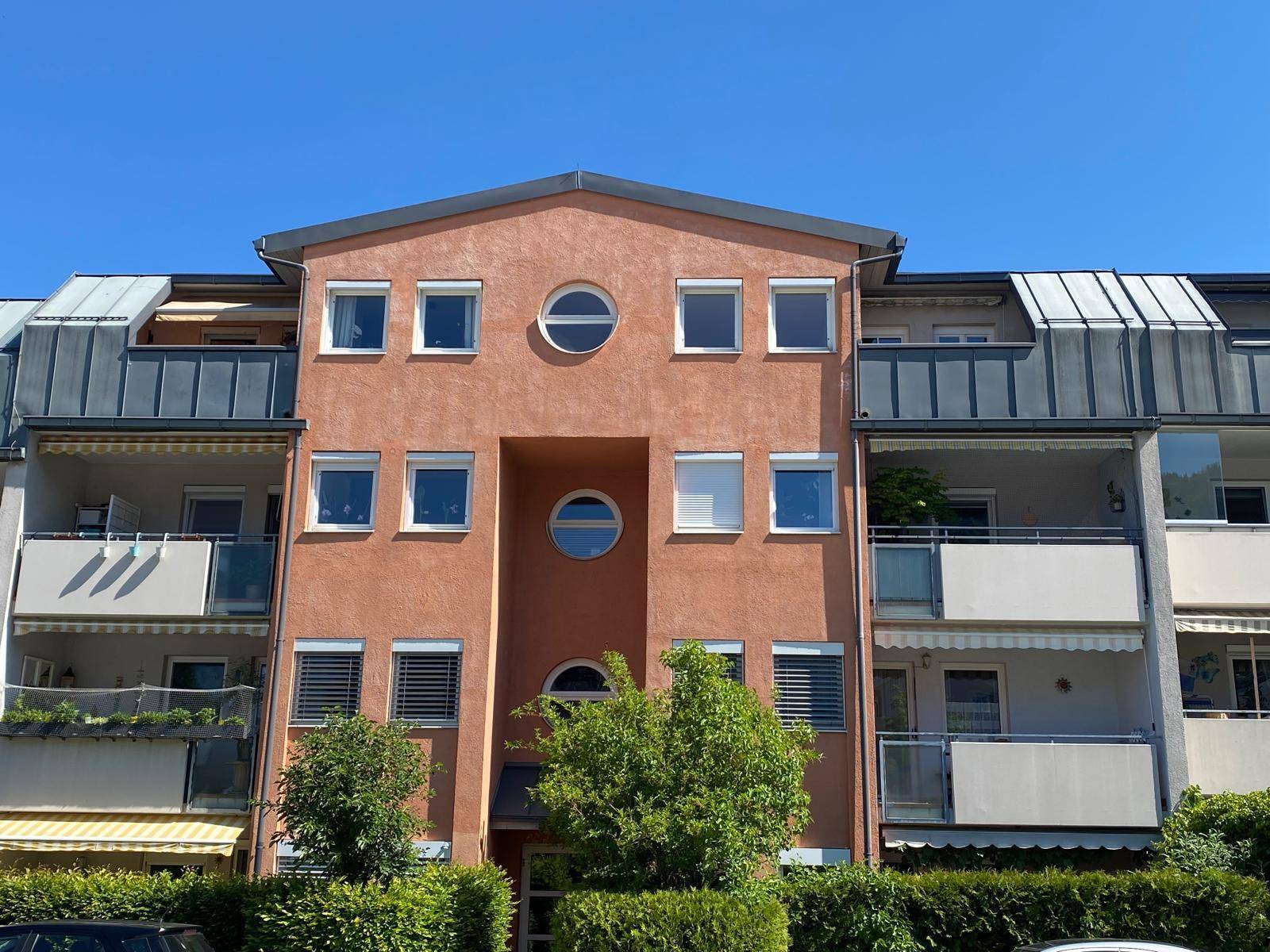 Moderne Gebäudefassade mit runden und rechteckigen Fenstern sowie Balkonen, umgeben von Bäumen.