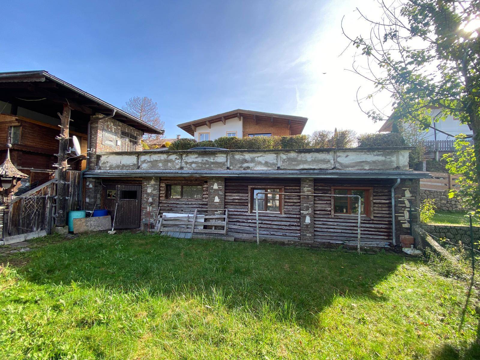 Rückseite des Hauses mit Anbau und Gartenbereich, umgeben von Bäumen unter blauem Himmel.