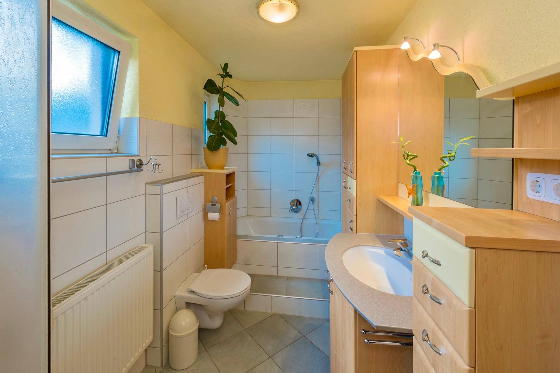 Helles Badezimmer mit Badewanne, WC, Waschbecken, Holzschränken und einem Fenster für Tageslicht.