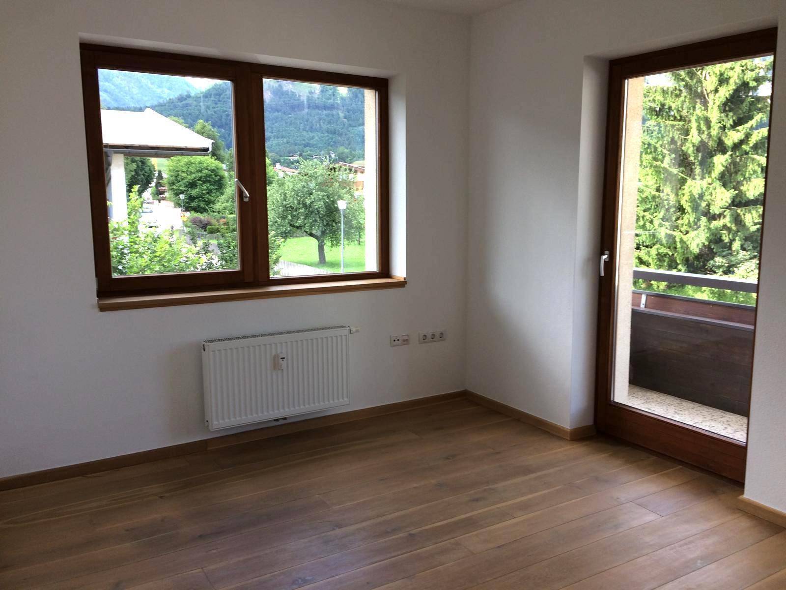 Leeres Zimmer mit Holzboden, großen Fenstern und Zugang zu einem Balkon.