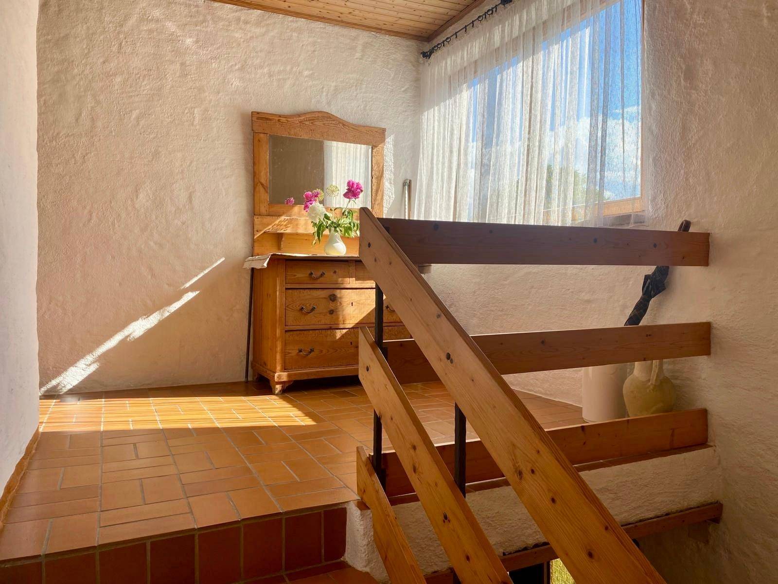 Heller Treppenaufgang mit Holzgeländer und Terrakottafliesen, beleuchtet durch ein großes Fenster.