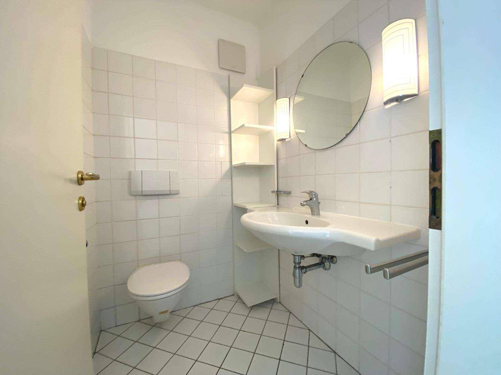Funktionales Badezimmer mit weißen Fliesen, Waschbecken und wandmontierter Toilette.