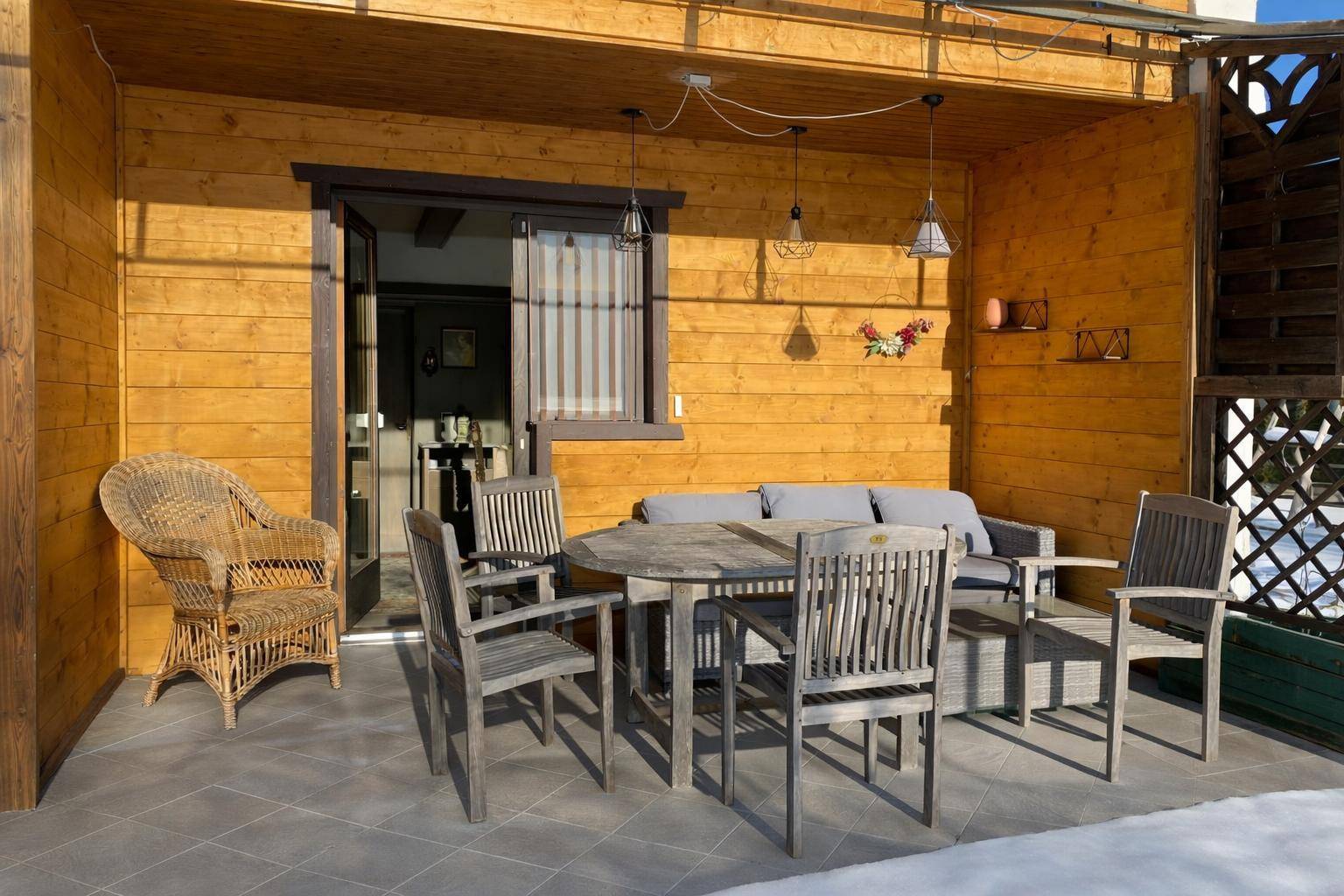 Überdachte Terrasse mit Holzwänden, Fliesenboden und rustikalen Sitzgelegenheiten im Freien.