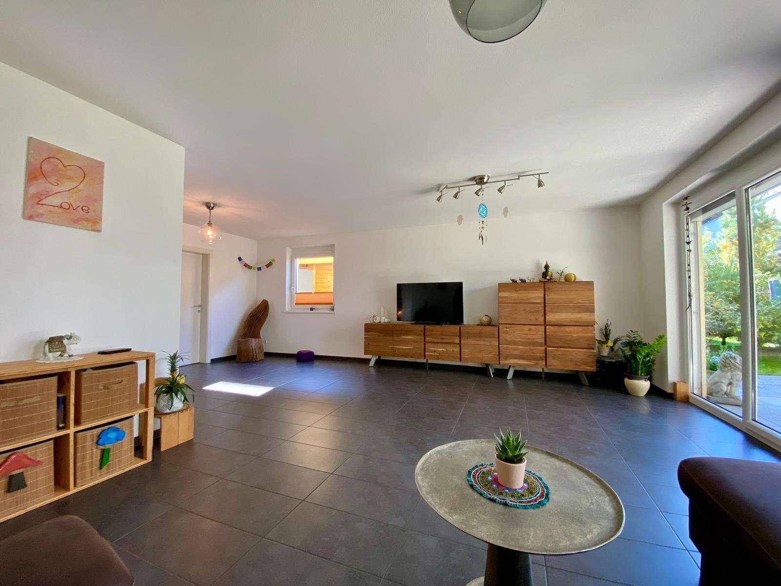 Geräumiges Wohnzimmer mit dunklen Fliesen, modernen Holzmöbeln und viel Tageslicht durch große Fenster.