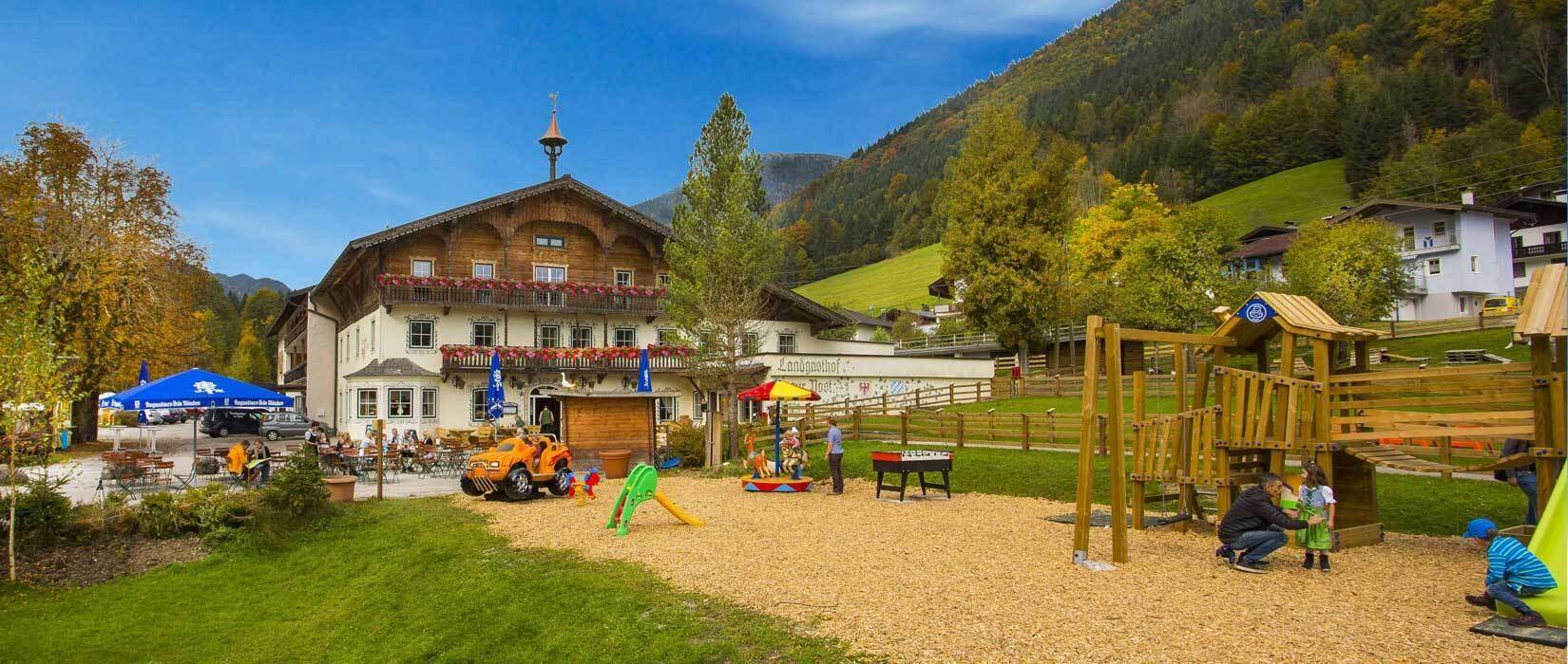 Weitläufiger Außenbereich mit großem Kinderspielplatz und dem traditionellen Gasthaus im Hintergrund, umgeben von Natur.