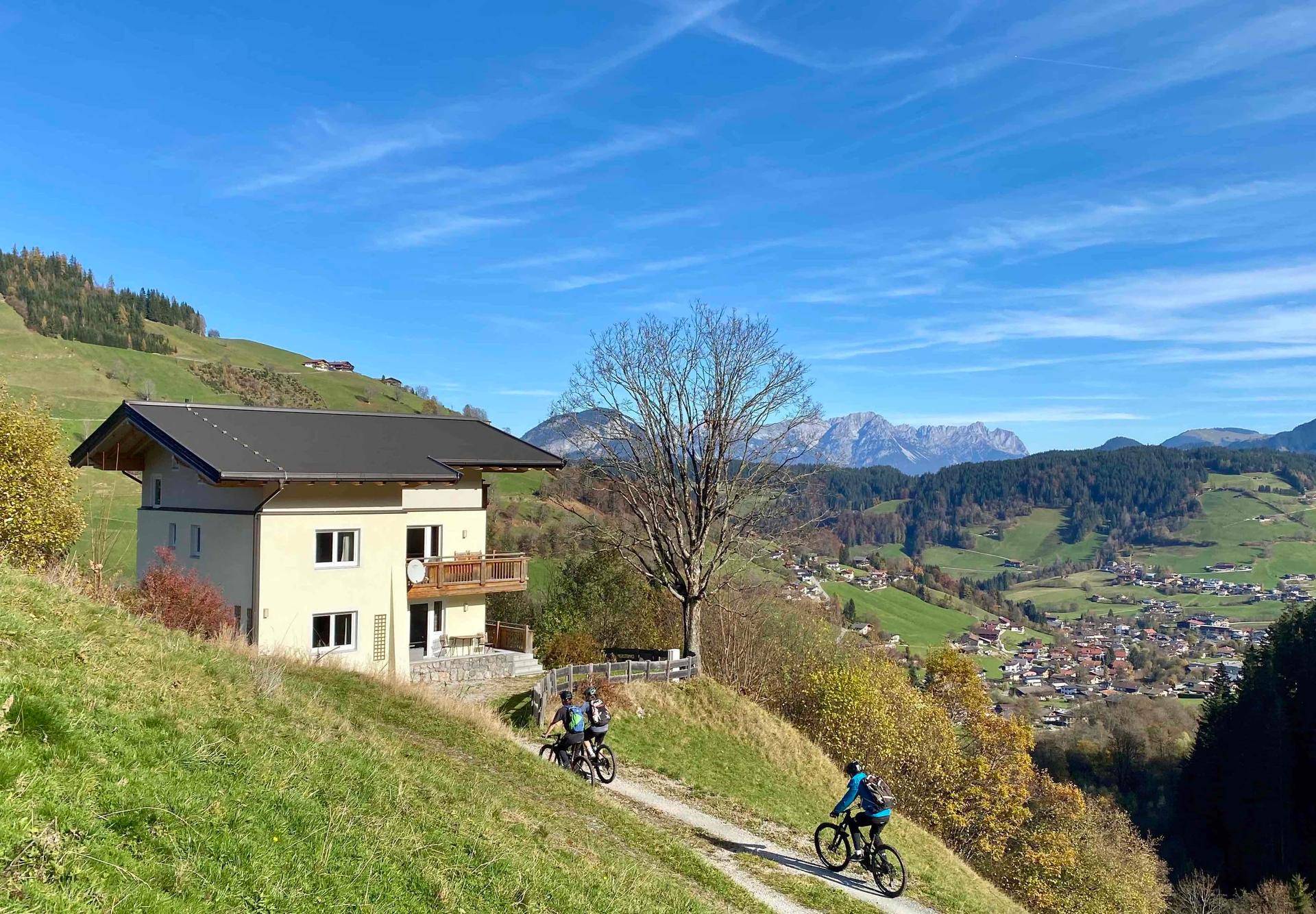 Das Haus in sonniger Hanglage mit Schotterweg und Radfahrern, umgeben von Berglandschaft.