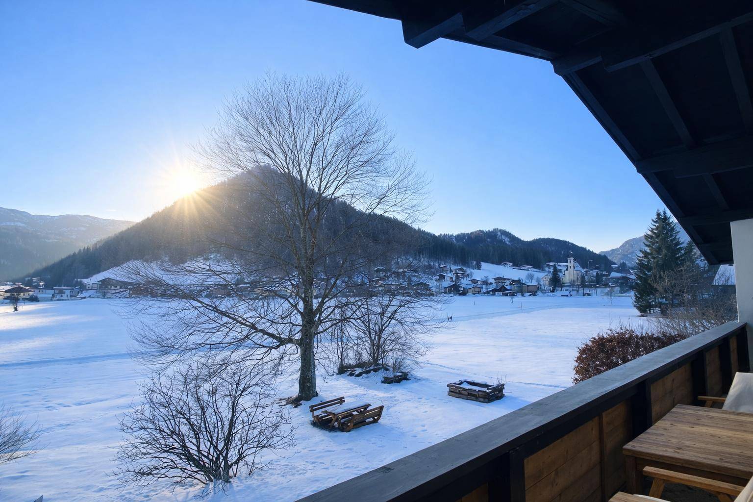 Sonniger Balkon mit Sitzgelegenheiten und weitem Blick auf die verschneite Landschaft und ein Dorf.