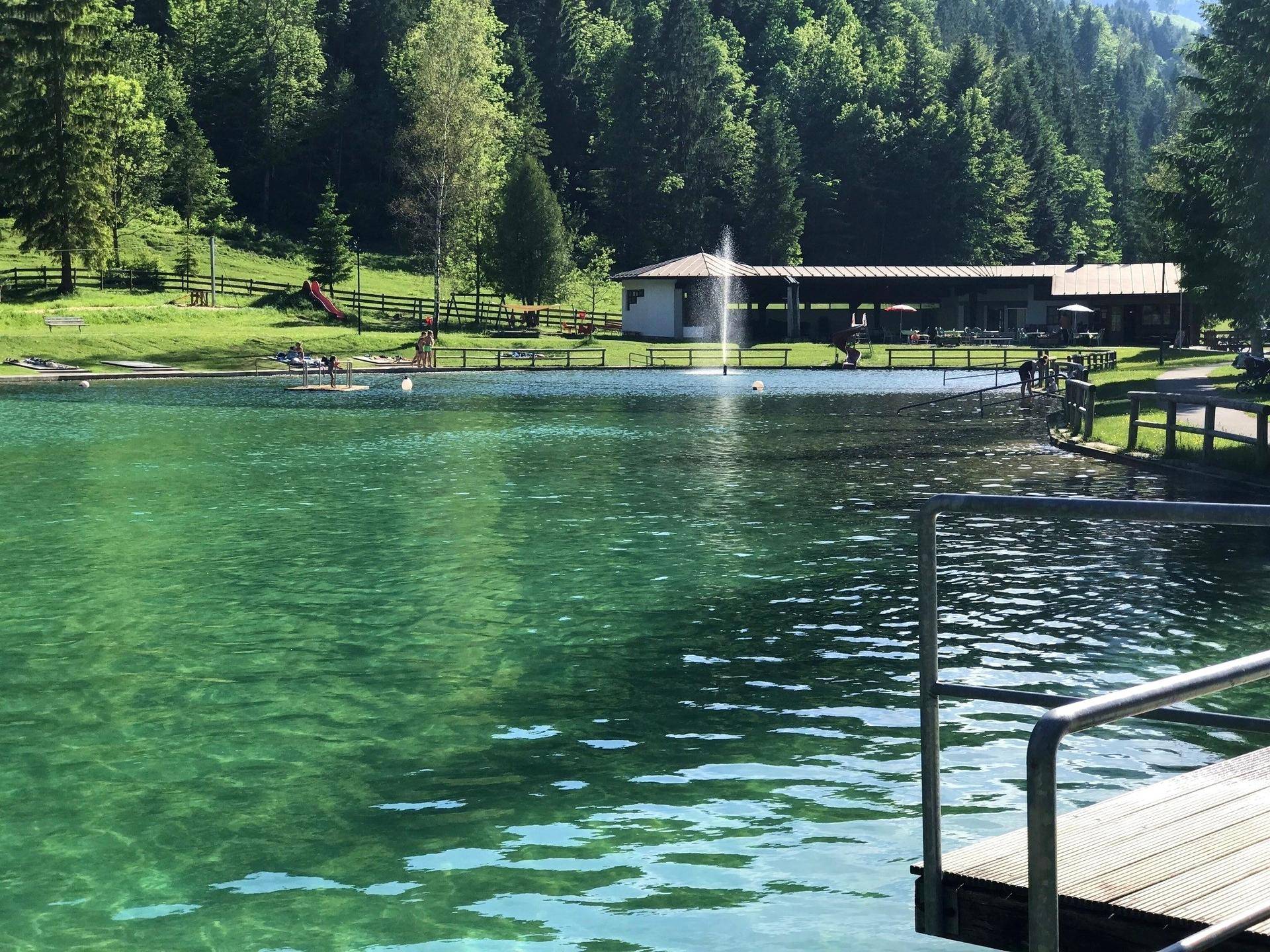 Klarer Badesee mit Fontäne und einem Gebäude im Hintergrund, umgeben von grünen Wäldern.