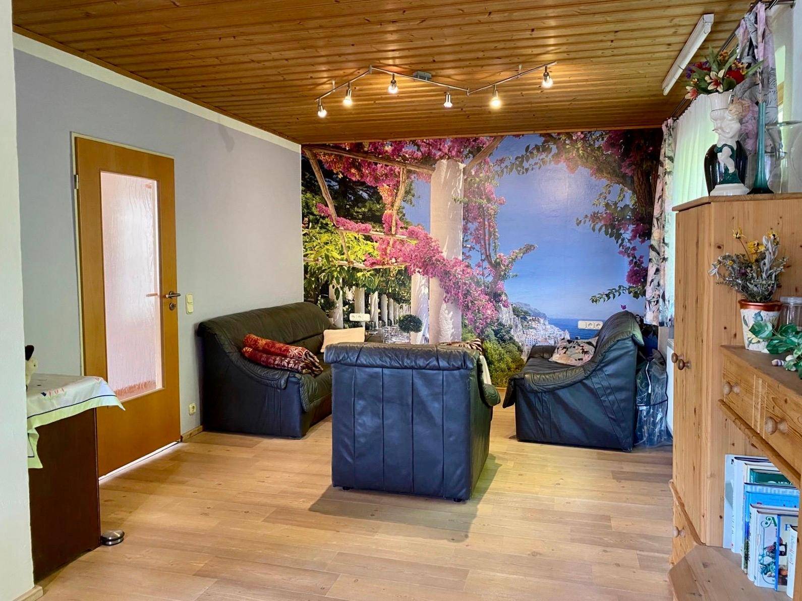 Gemütliches Wohnzimmer mit Ledersofas, Holzdecke und einer großen Wandtapete mit Landschaftsmotiv.