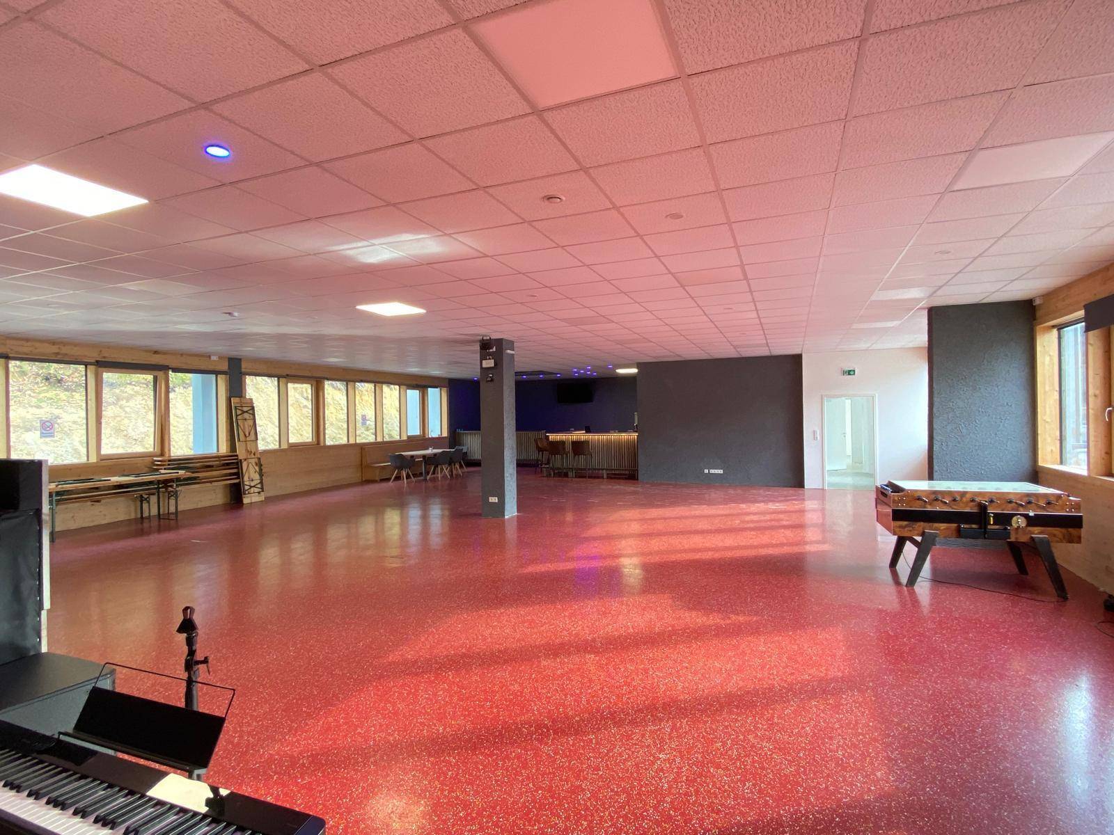 Großer Mehrzweckraum mit rotem Boden, vielen Fenstern und einer Bar im Hintergrund, ideal für Veranstaltungen.