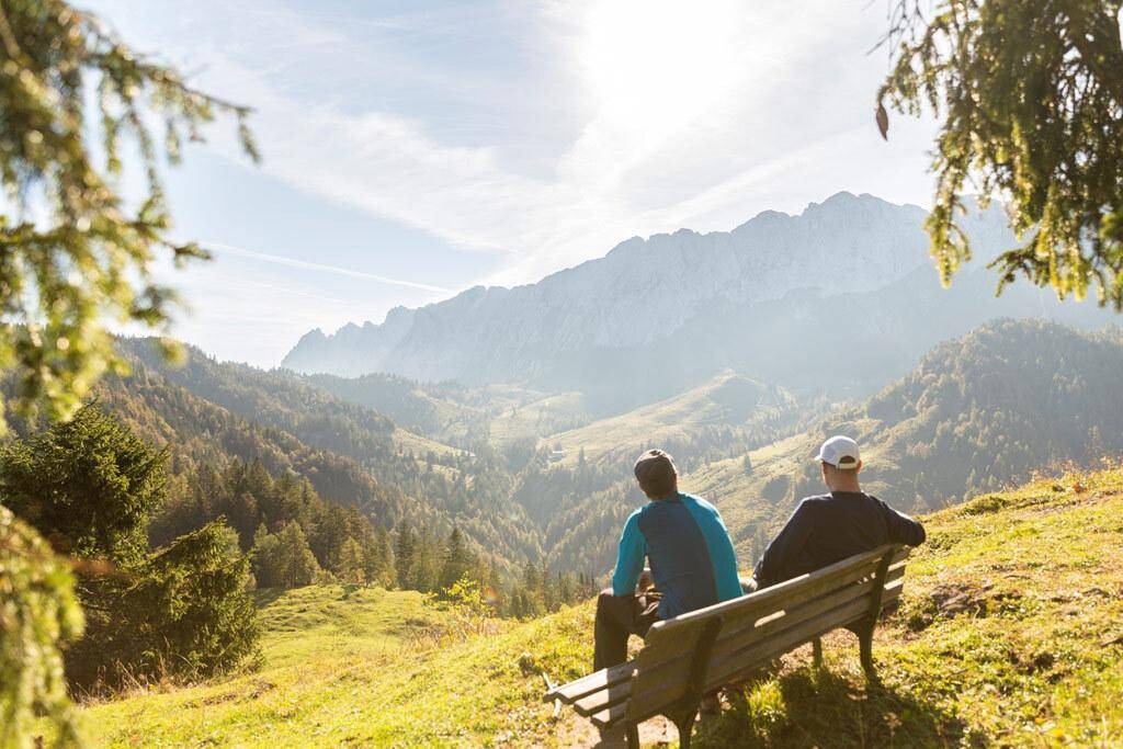 Zwei Personen genießen die weite Aussicht auf die Berge und Täler von einer Bank aus.