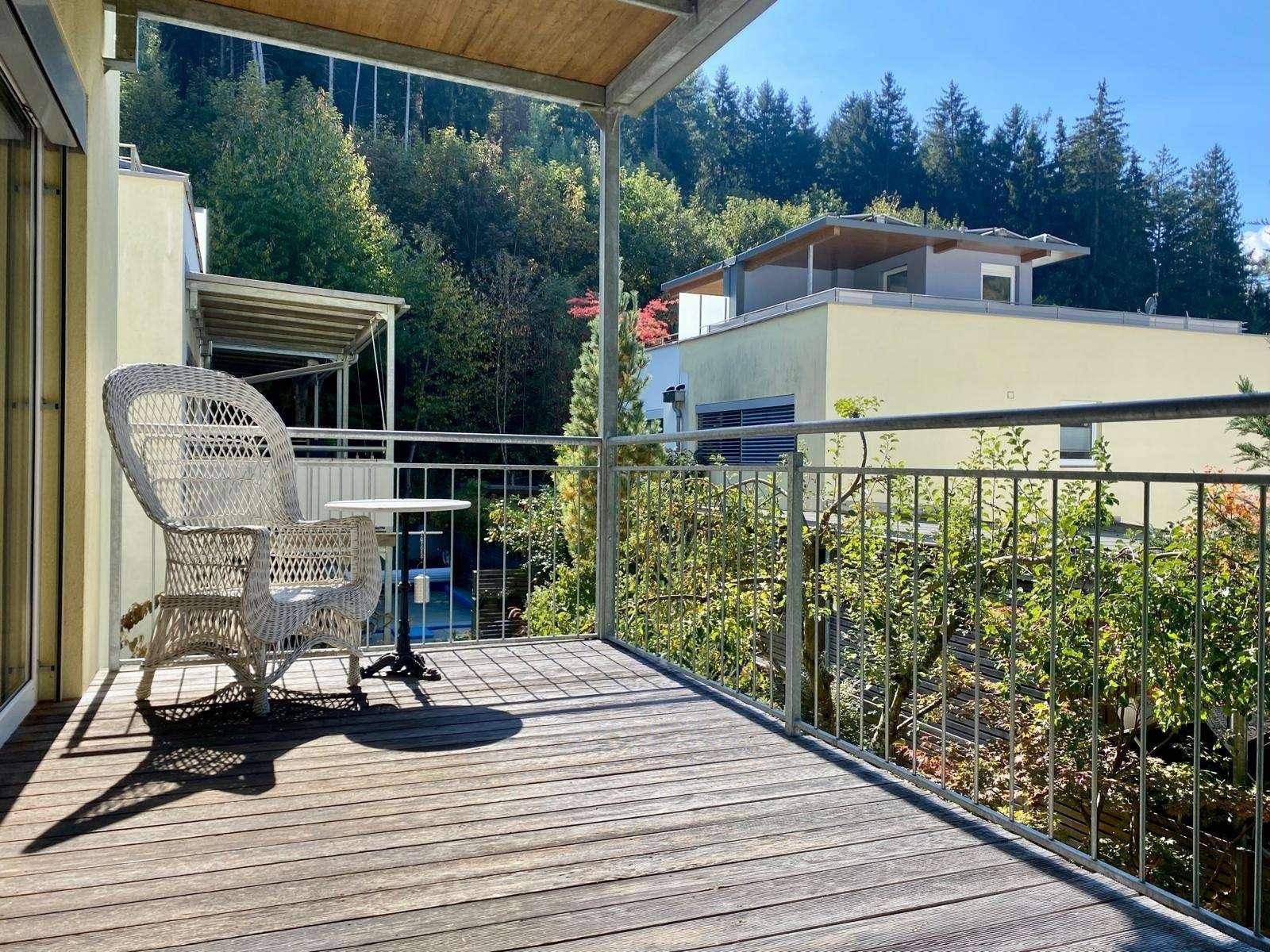 Geräumiger Holzbalkon mit Korbsessel und Blick auf die umliegende grüne Landschaft unter blauem Himmel.