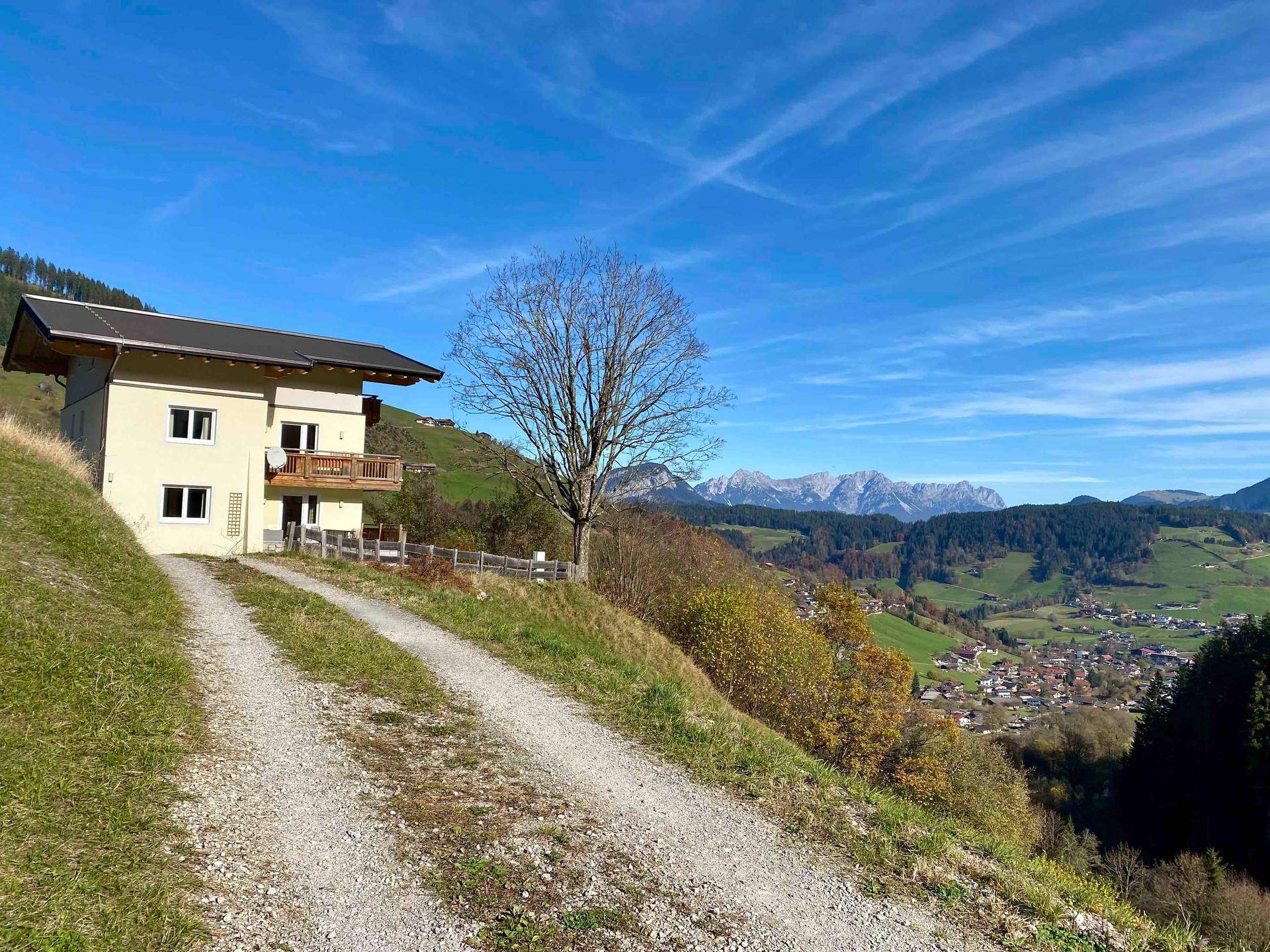 Das Haus in sonniger Hanglage mit Schotterweg und weitem Blick auf die Bergwelt.