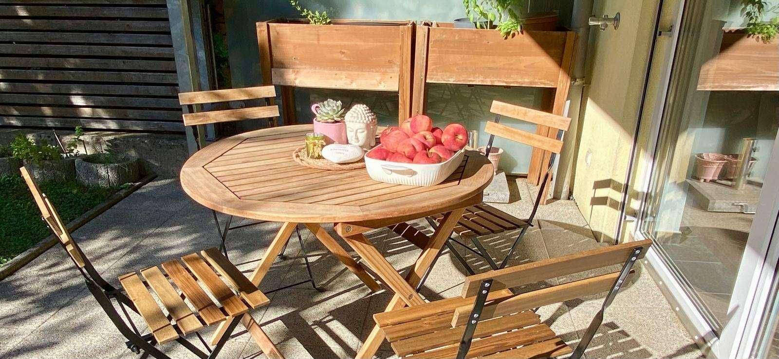 Gemütliche Terrasse mit Holztisch und Stühlen, umgeben von Pflanzen in Holzkästen unter Sonnenschein.