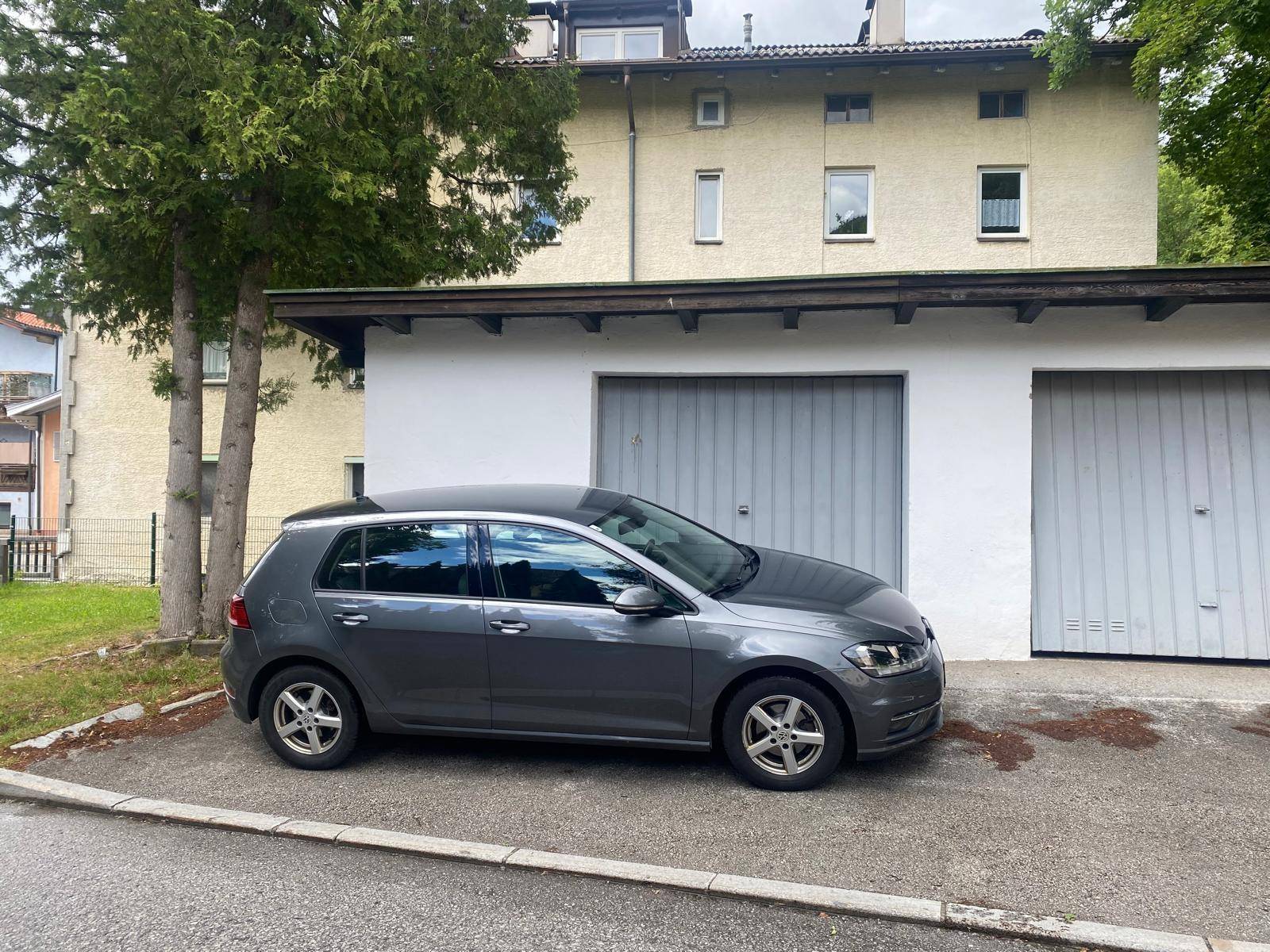 Grauer VW Golf parkt vor einem Garagengebäude mit dahinterliegendem Wohnhaus und Bäumen.