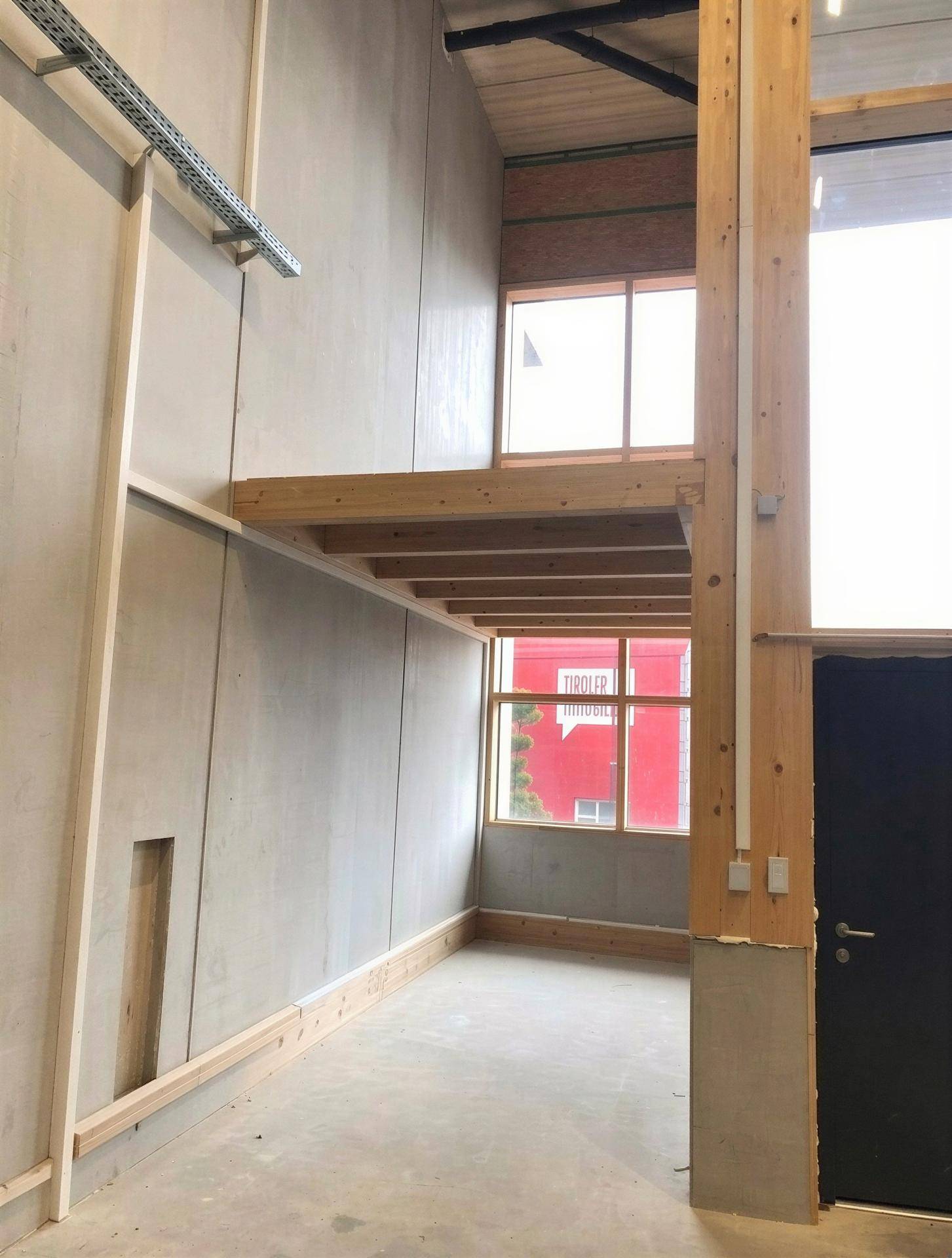 Hoher Gewerberaum mit Betonwänden, einer Holzempore und großen Fenstern, die viel Tageslicht hereinlassen.