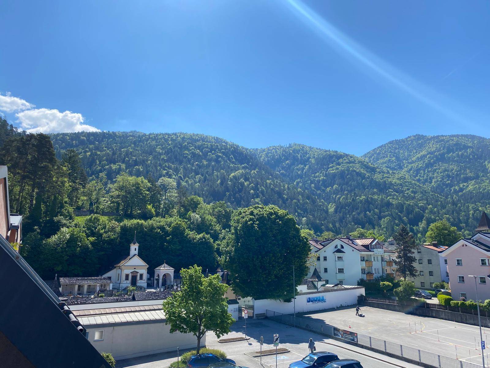 Panoramablick auf ein grünes Tal mit bewaldeten Bergen, Häusern und einer kleinen Kirche unter blauem Himmel.