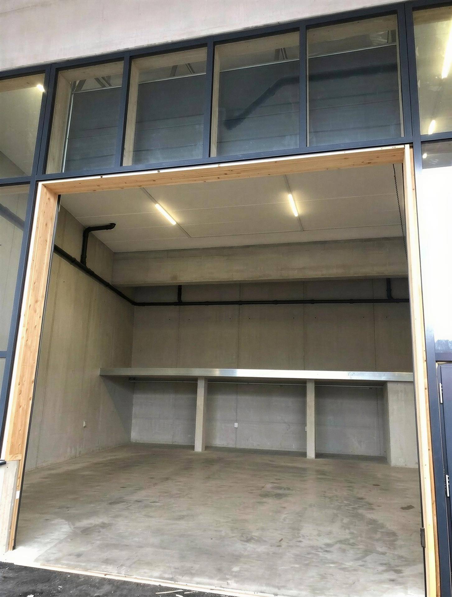 Leerer Gewerberaum mit hohen Decken, Betonboden und einer großen Eingangstür mit Fensterfront.