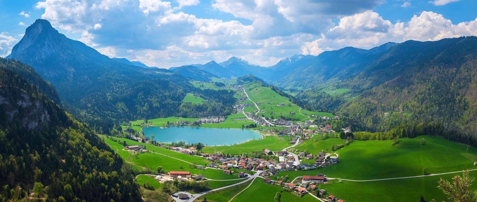 Malerische Alpenlandschaft mit einem See, einem Dorf und grünen Tälern unter blauem Himmel.