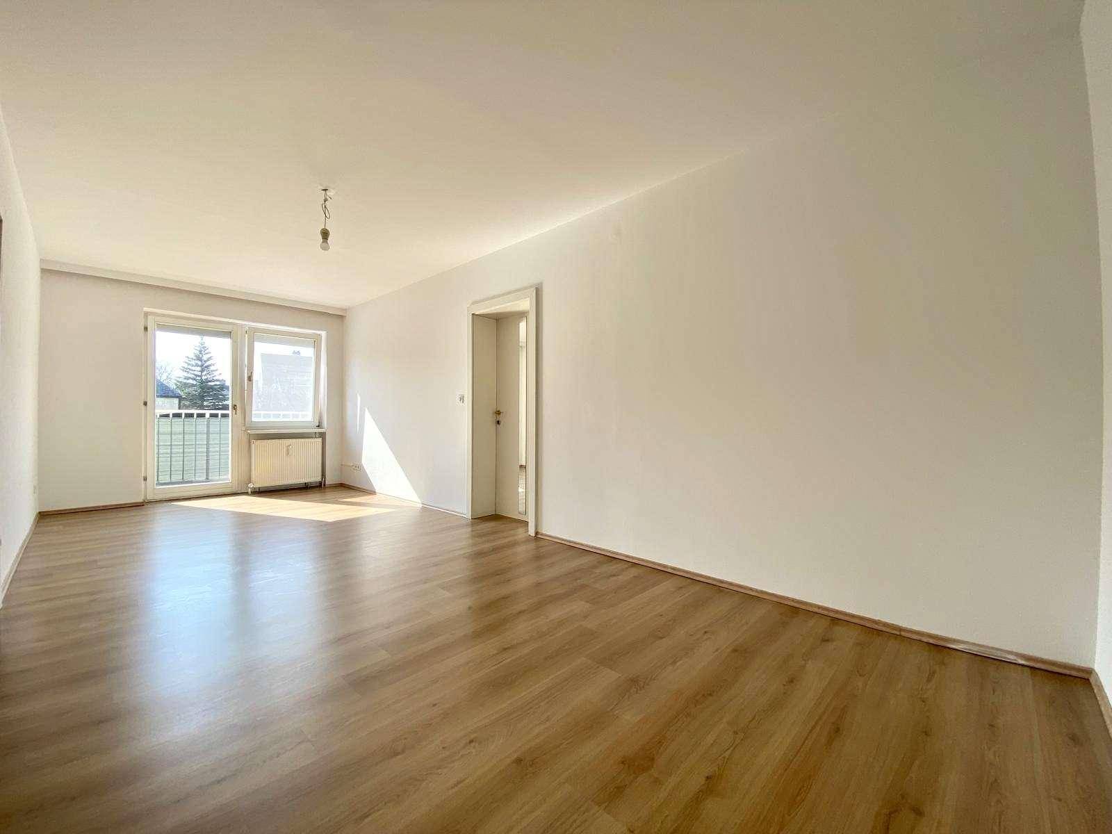 Helles, leeres Wohnzimmer mit Laminatboden und Zugang zu einem Balkon.