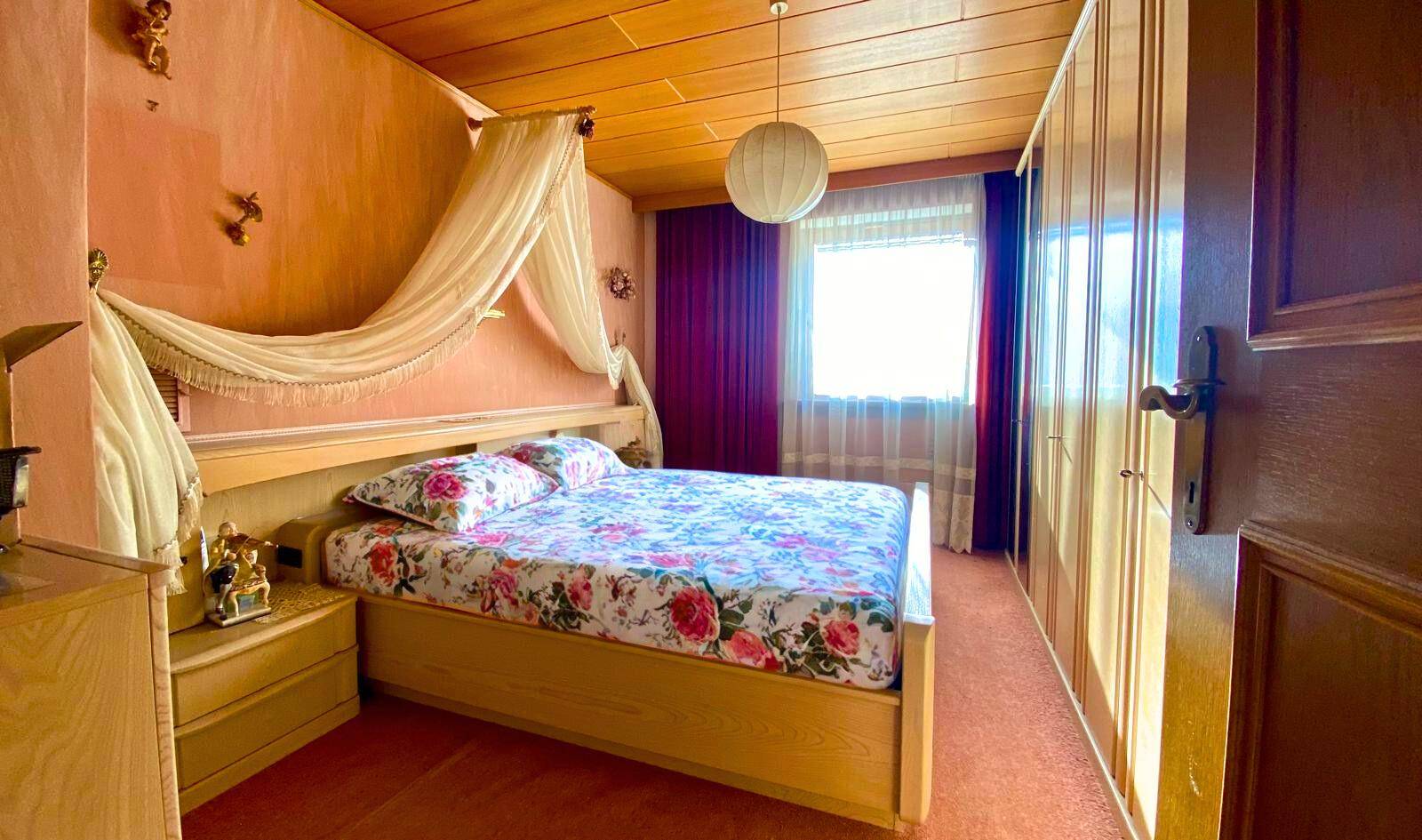 Schlafzimmer mit Doppelbett, Holzdecke, Kleiderschrank und Fenster mit Vorhängen.