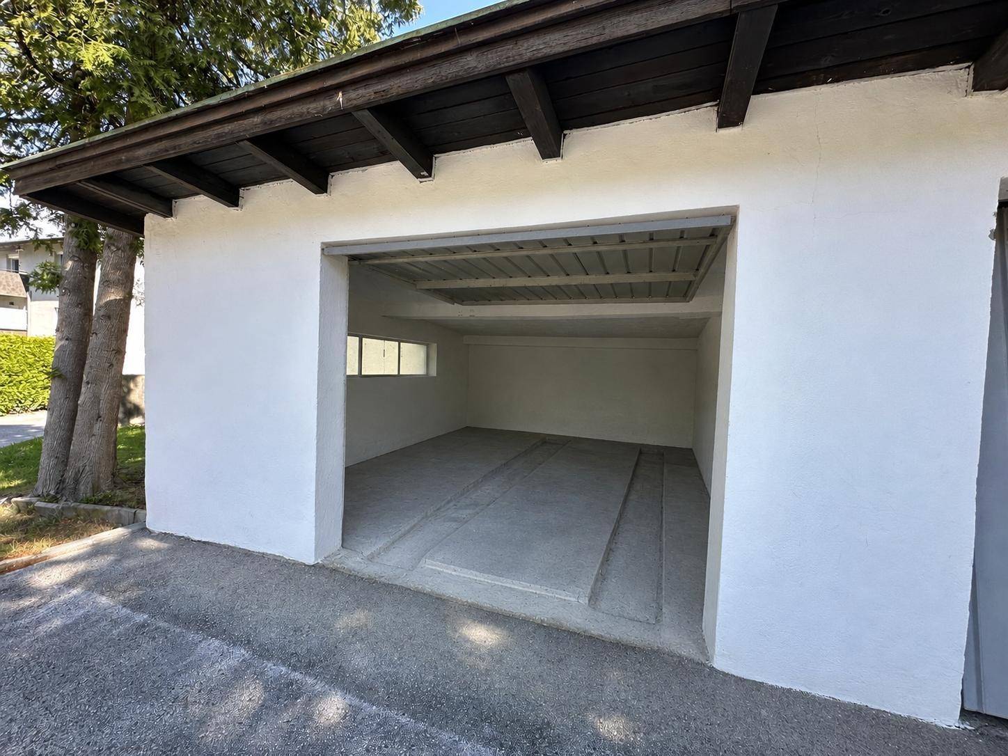 Offene Garagenbox mit Blick auf den Betonboden und das kleine Fenster, umgeben von Bäumen.