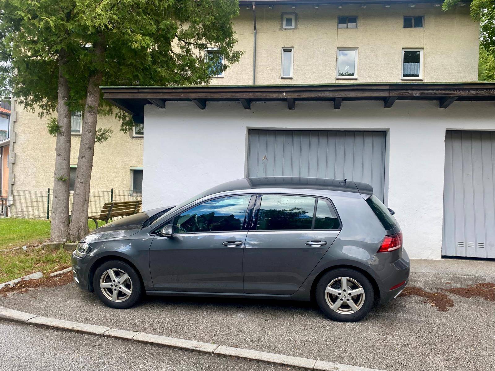 Ansicht des grauen VW Golfs vor den Garagen und dem dahinterliegenden Wohngebäude mit Grünflächen.
