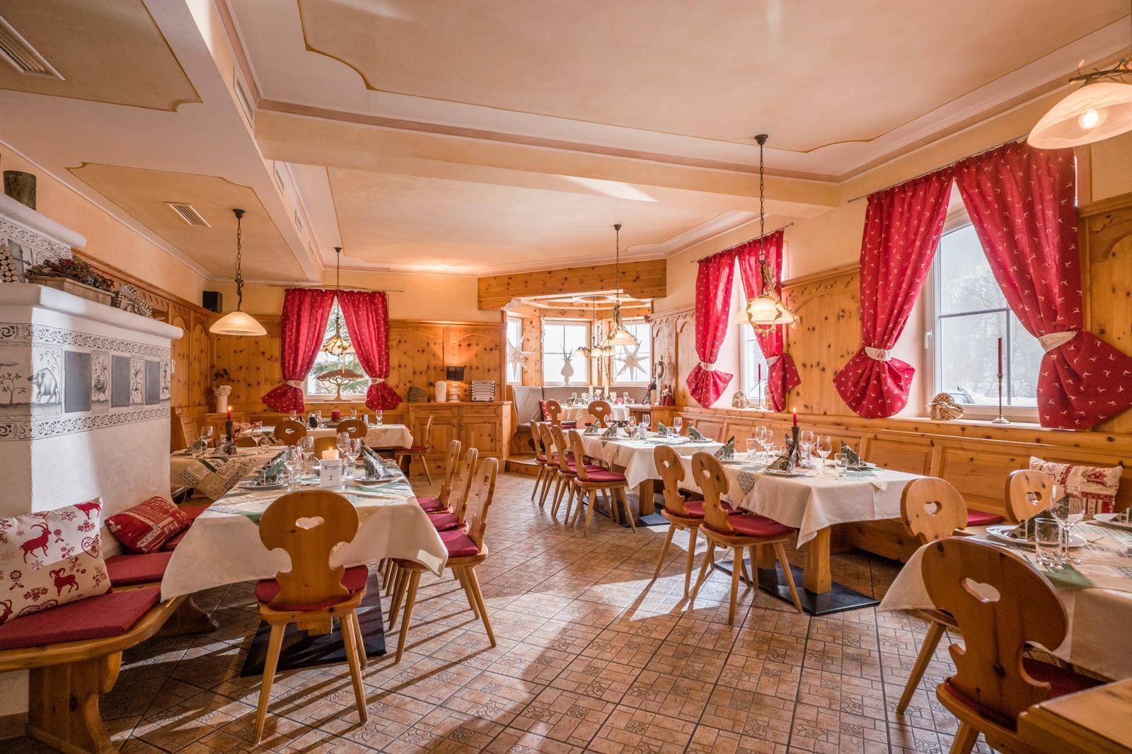 Gemütlicher Speisesaal im traditionellen Stil mit Holztäfelung, Kachelofen und roten Vorhängen.