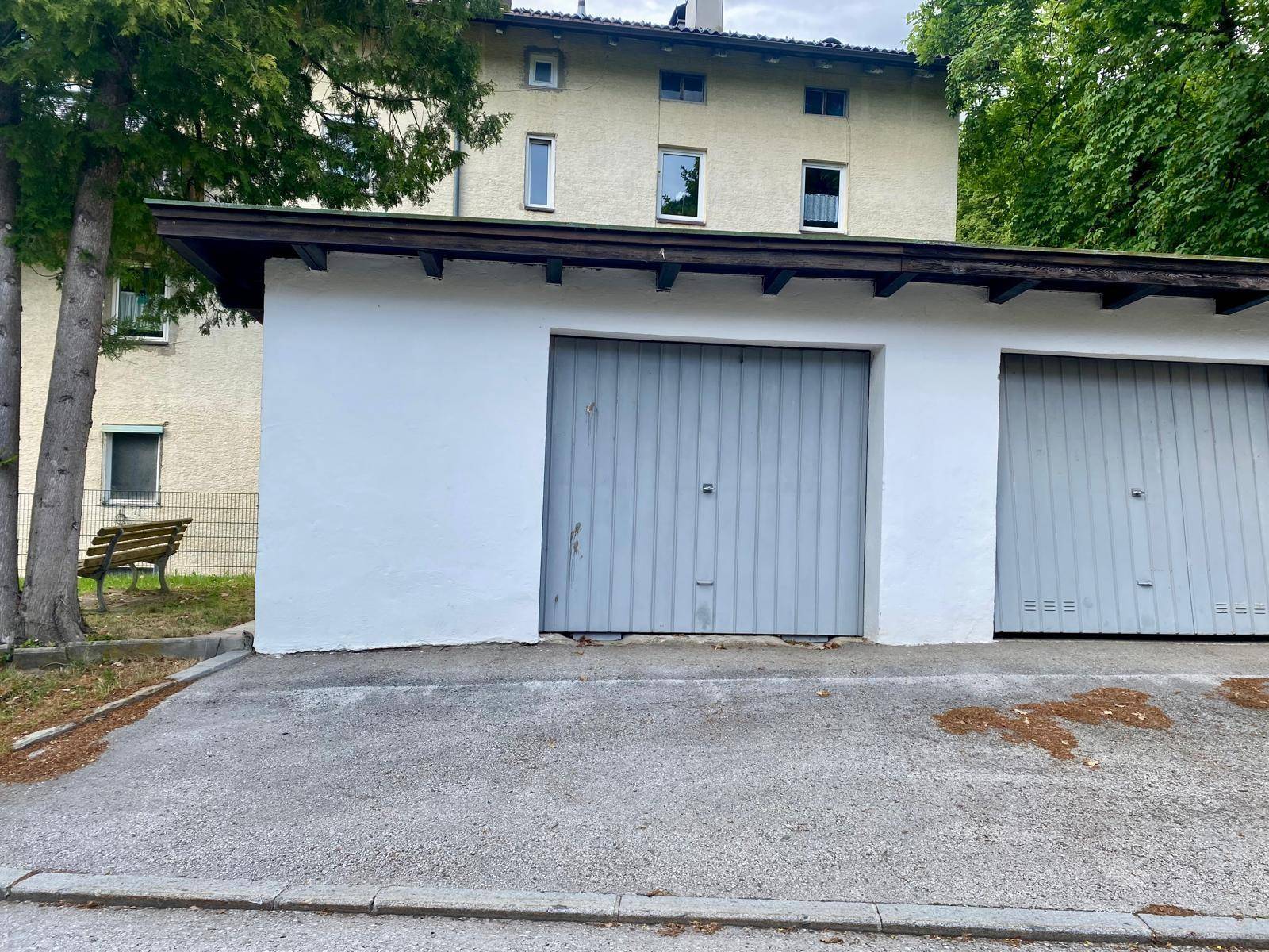 Mehrere Garagenboxen mit grauen Toren und weißer Fassade, im Hintergrund ein Wohngebäude mit Bäumen.