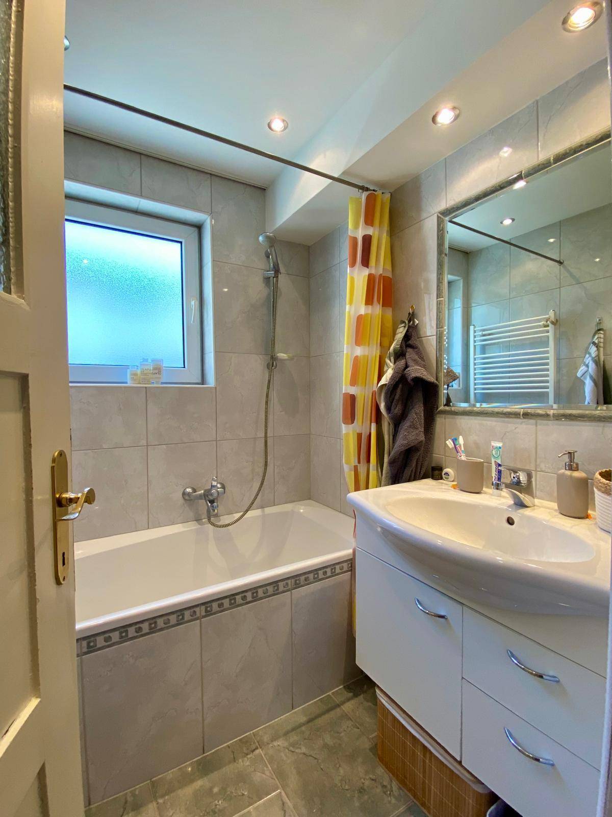 Badezimmer mit Badewanne, Waschbecken und Fenster, hell gefliest.