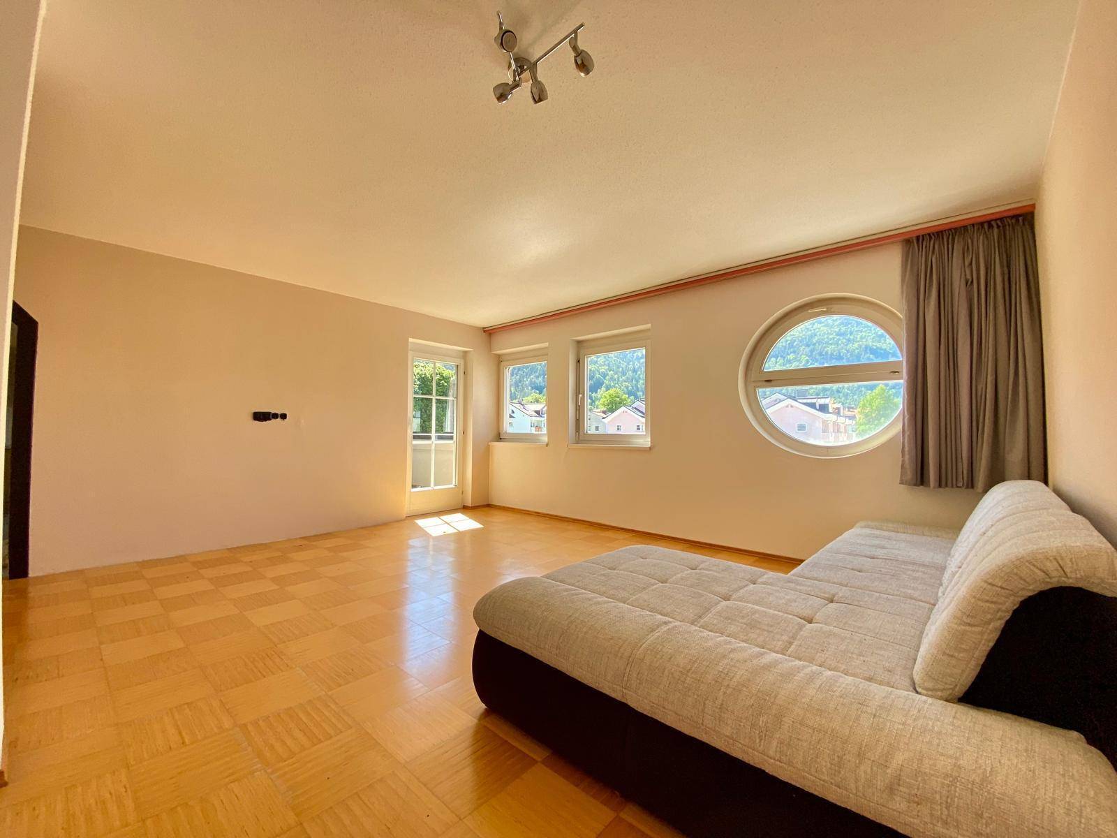 Helles Wohnzimmer mit Parkettboden, einem Sofa und verschiedenen Fenstern, darunter ein Rundfenster.
