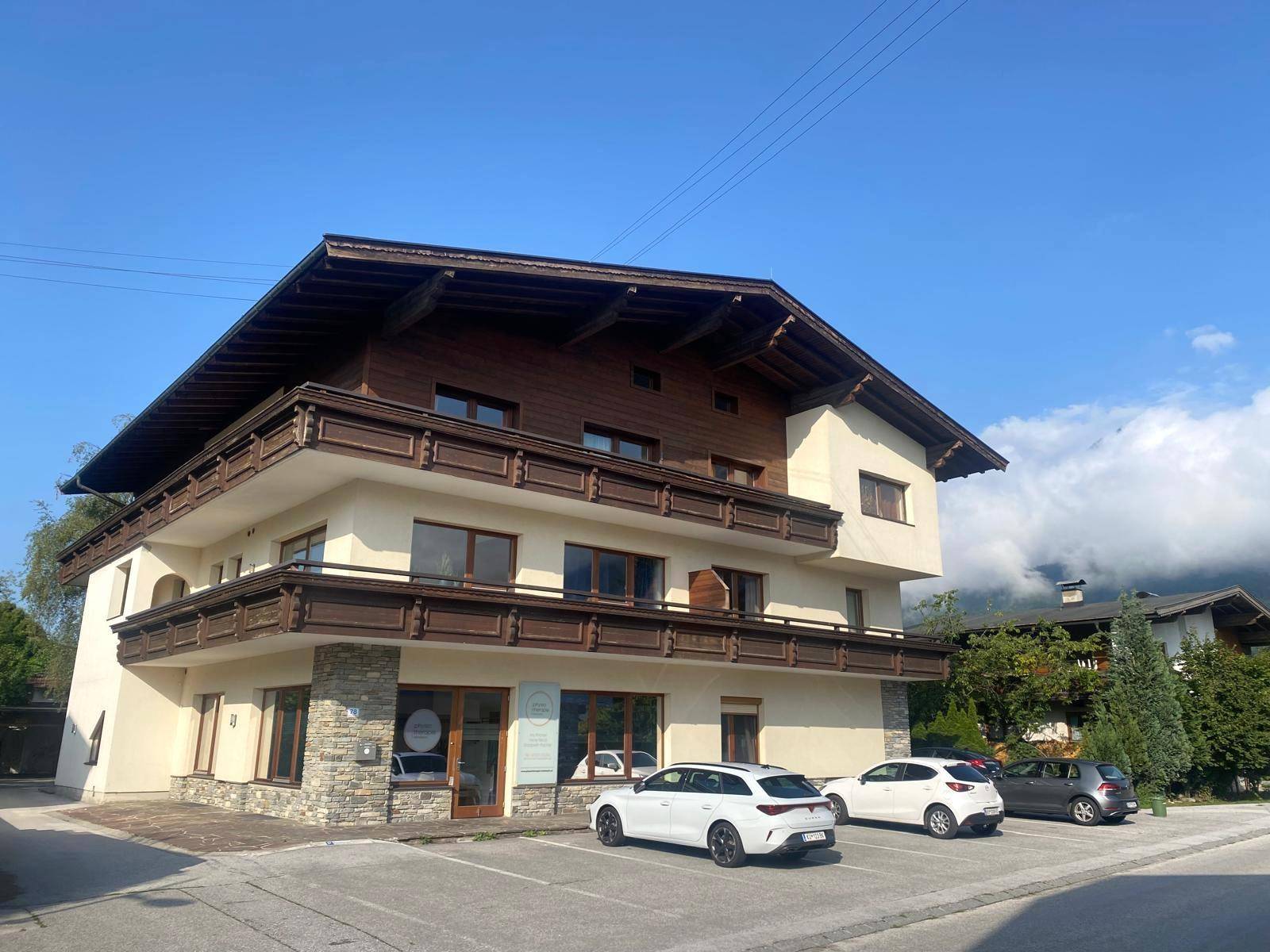 Gebäude im alpinen Stil mit Holzbalkonen, Steinverkleidung und Parkplätzen unter blauem Himmel.