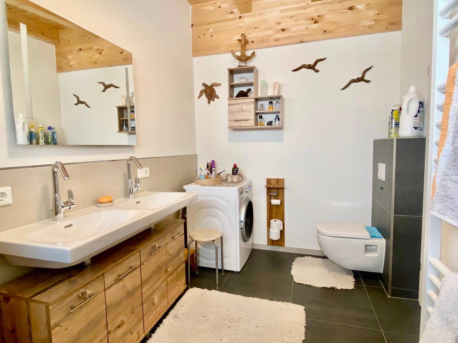 Modernes Badezimmer mit Doppelwaschbecken, Wand-WC und Waschmaschine, unter Holzbalkendecke.
