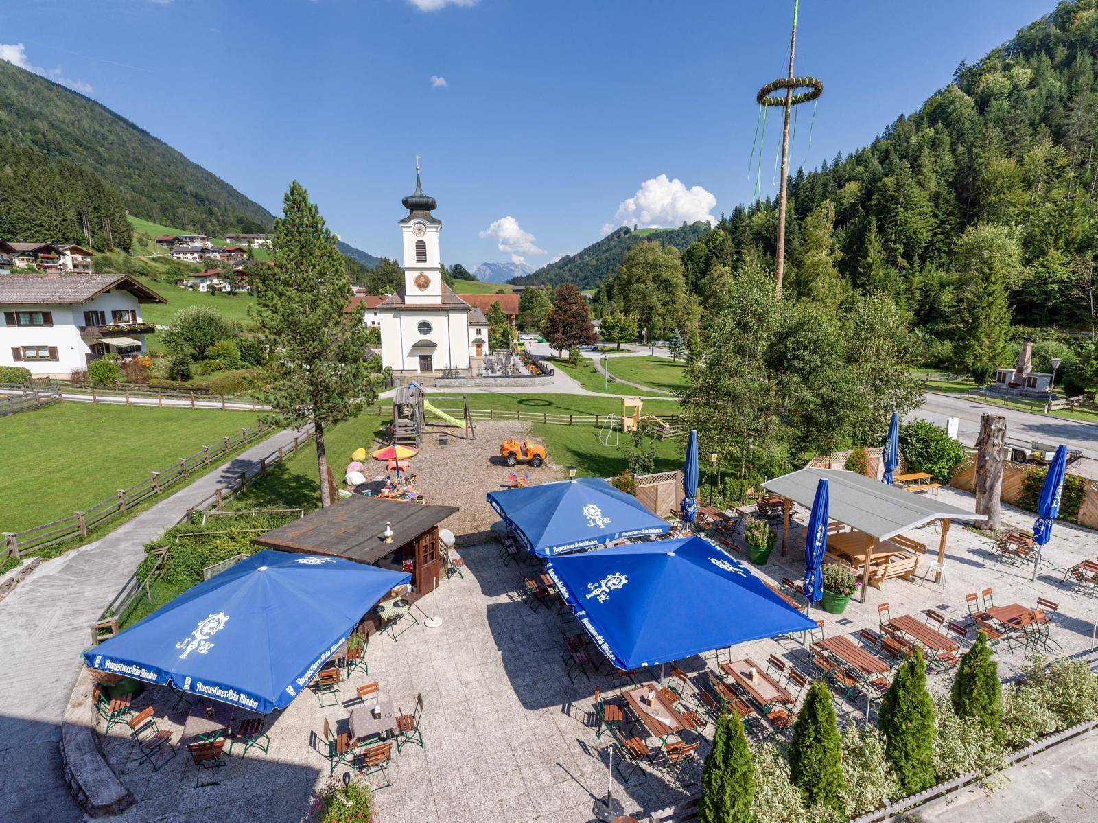 Großer Außenbereich mit Sonnenschirmen, Tischen und einem Kinderspielplatz vor einer malerischen Bergkulisse.