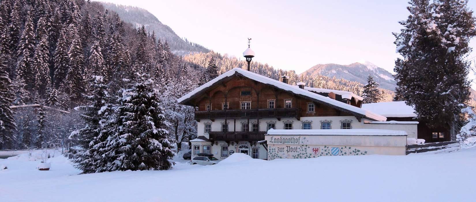 Traditionelles Gasthaus in einer verschneiten Winterlandschaft, umgeben von schneebedeckten Bäumen und Bergen.
