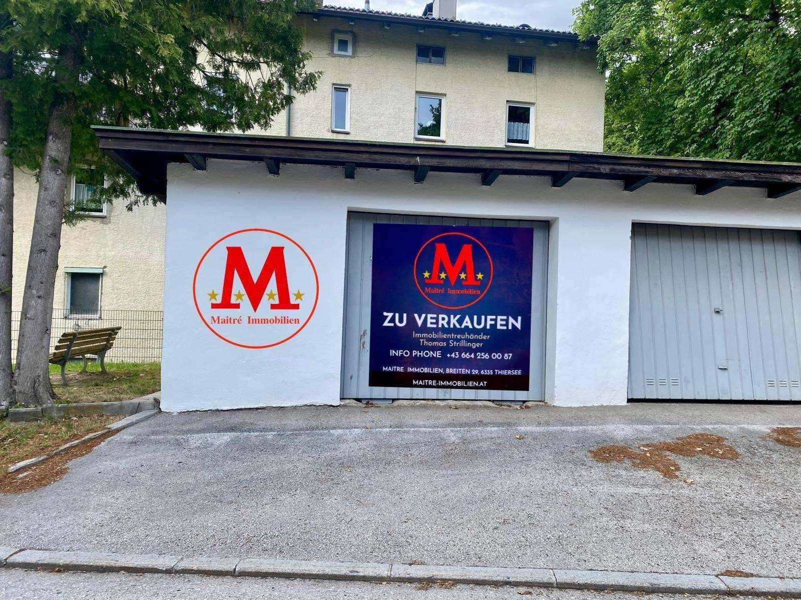 Außenansicht der Garage mit einem "ZU VERKAUFEN"-Banner und dem dahinterliegenden Wohngebäude.