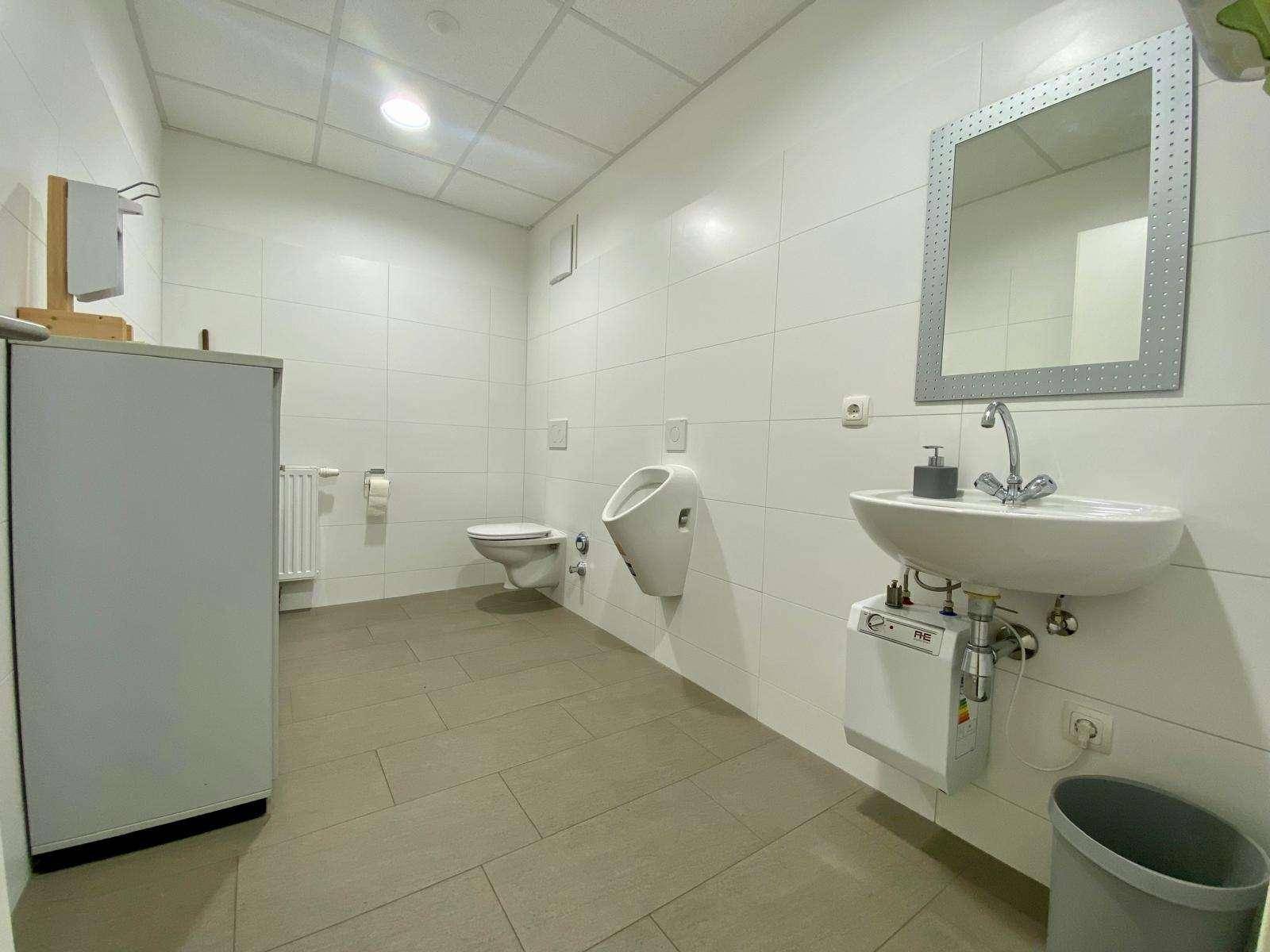 Modernes Badezimmer mit weißen Fliesen, ausgestattet mit Toilette, Urinal und einem Waschbecken mit Spiegel.