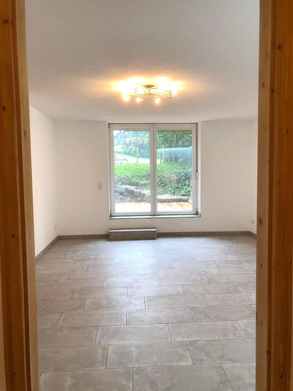 Helles, leeres Zimmer mit großen Fliesen und einem Fenster mit Blick ins Grüne.