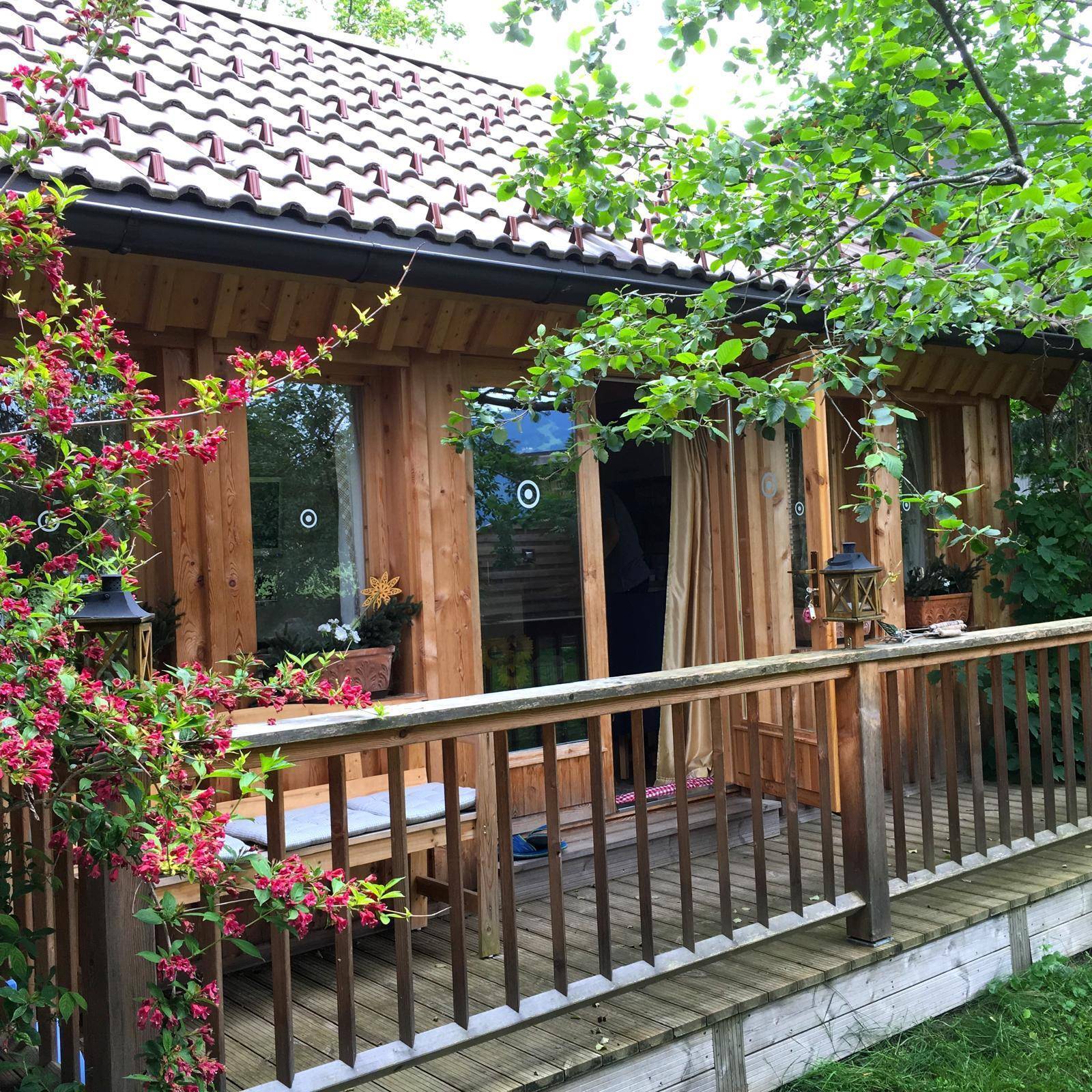 Detailansicht des Holzhauses mit Holzterrasse und blühenden Pflanzen, umgeben von Natur.