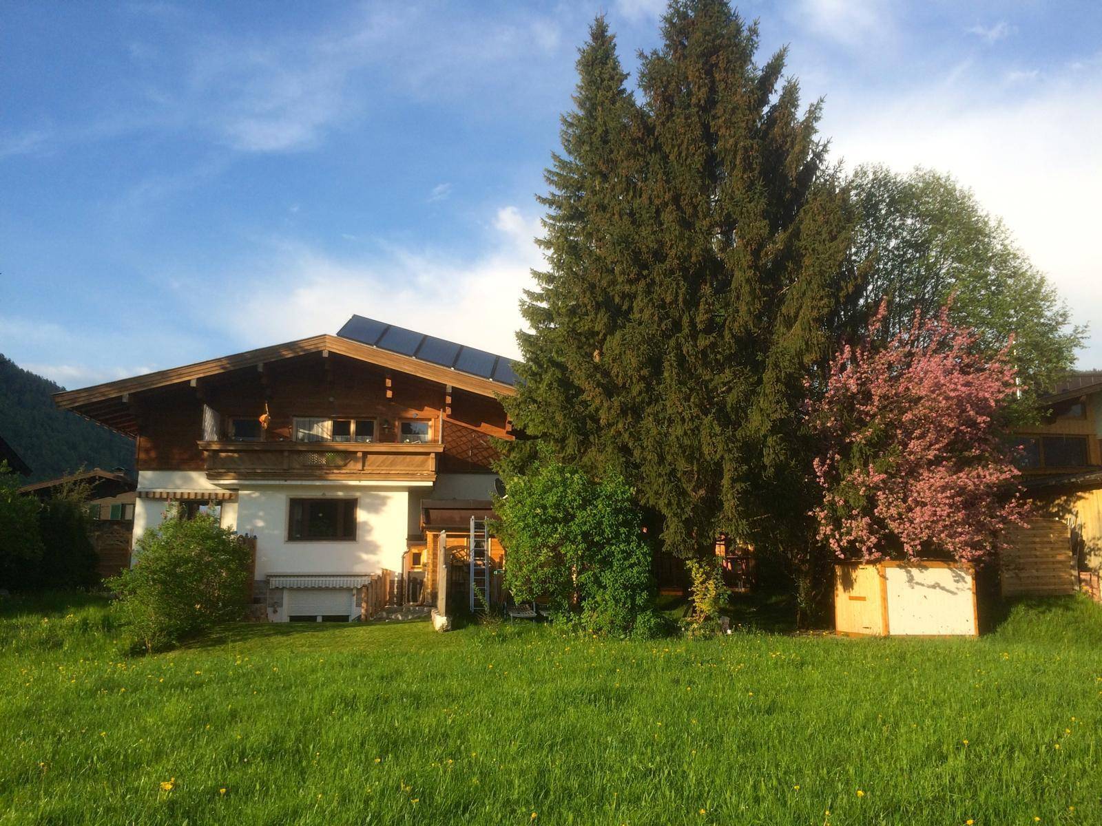 Alpenländisches Haus mit Solaranlage und blühendem Baum im Vorgarten bei Sonnenuntergang.