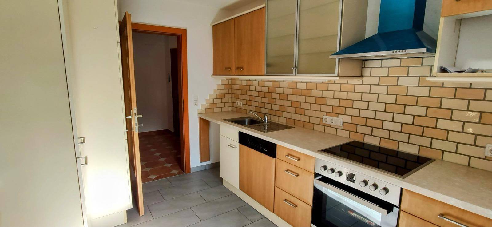 Funktionale Küche mit Holzfronten, Fliesenrückwand, Spüle, Herd und blauer Dunstabzugshaube.