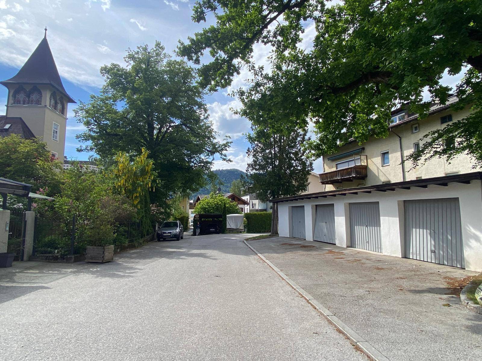 Wohnstraße mit Garagenreihe, Bäumen und einem Kirchturm im Hintergrund unter bewölktem Himmel.