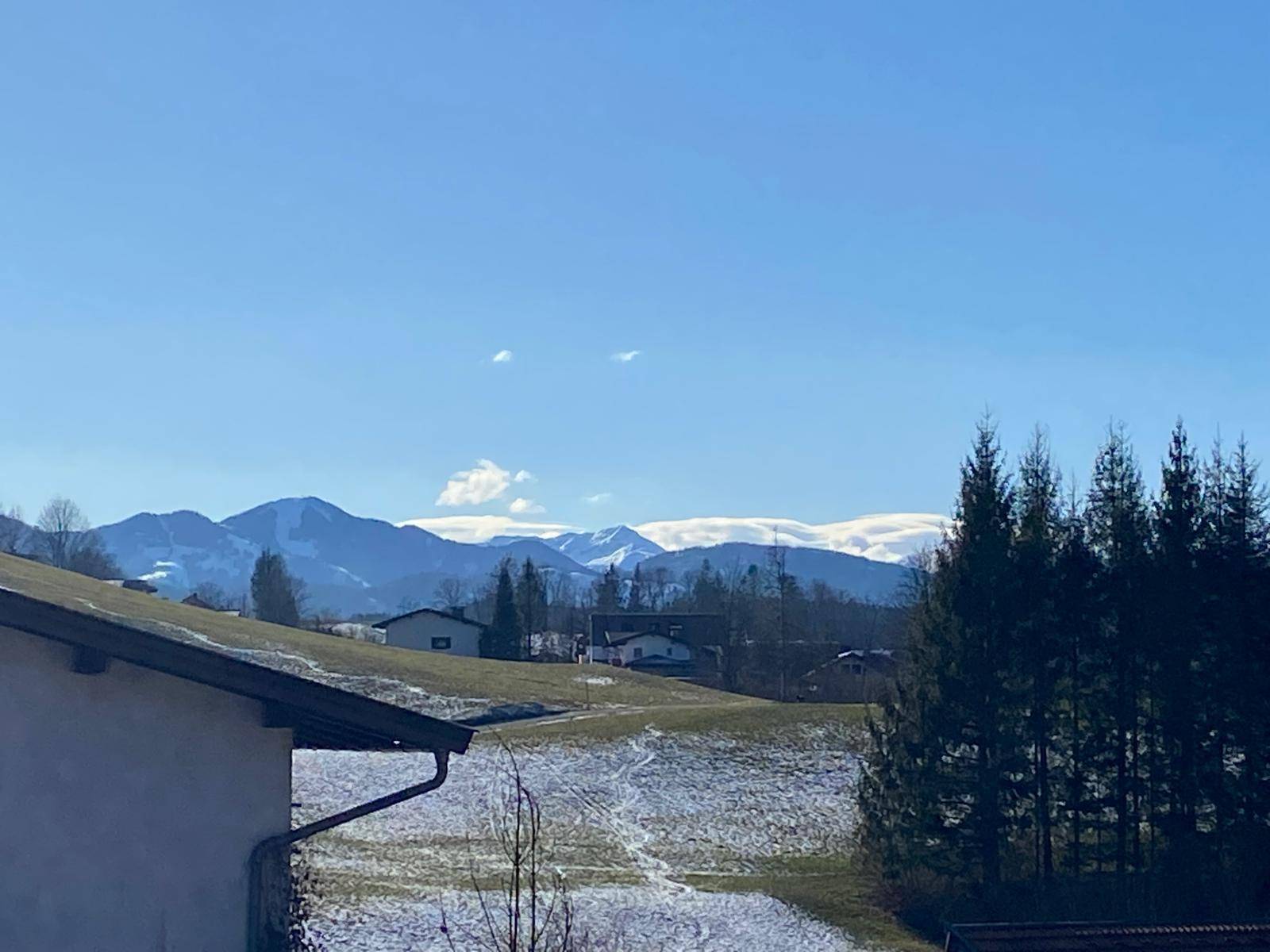 Blick auf schneebedeckte Berggipfel in der Ferne über einem Hausdach und grünen Hügeln.