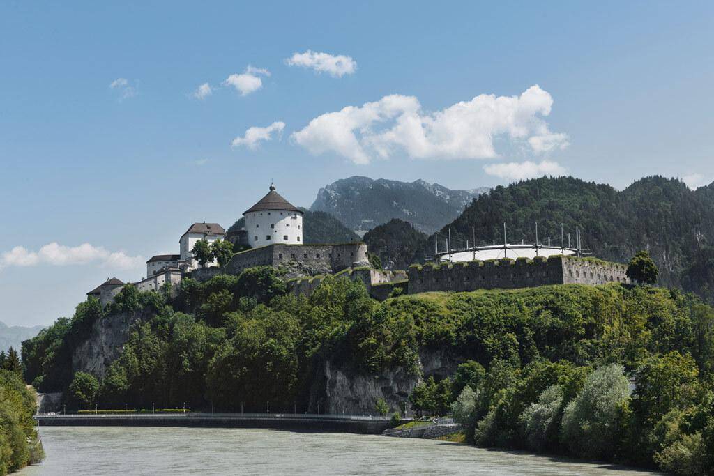 Historische Festung auf einem Hügel über einem Fluss, umgeben von grünen Bäumen und Bergen.