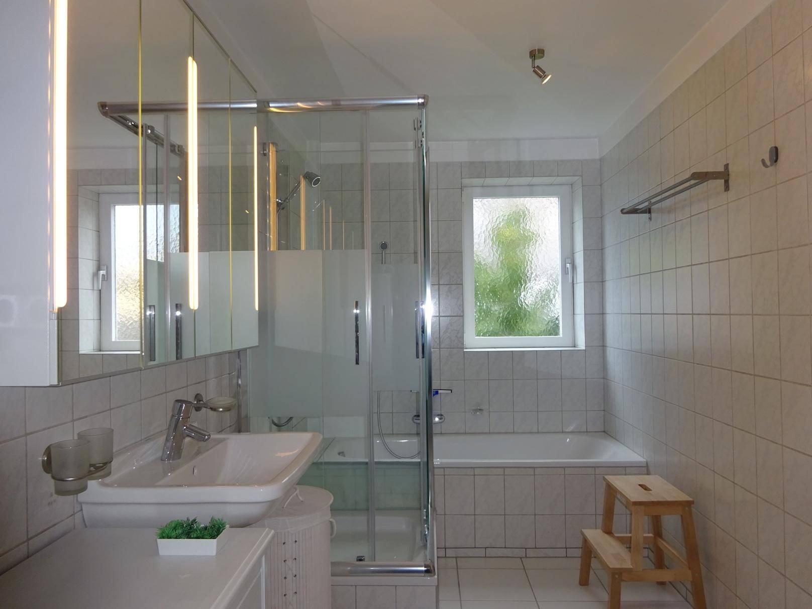 Helles Badezimmer mit weißen Fliesen, Badewanne, Duschkabine und Fenster.