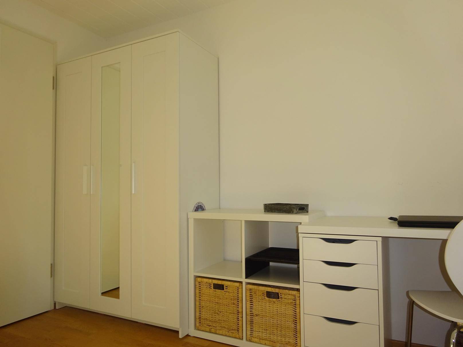 Weißer Kleiderschrank und Schreibtisch mit Ablageflächen in einem hellen Zimmer.