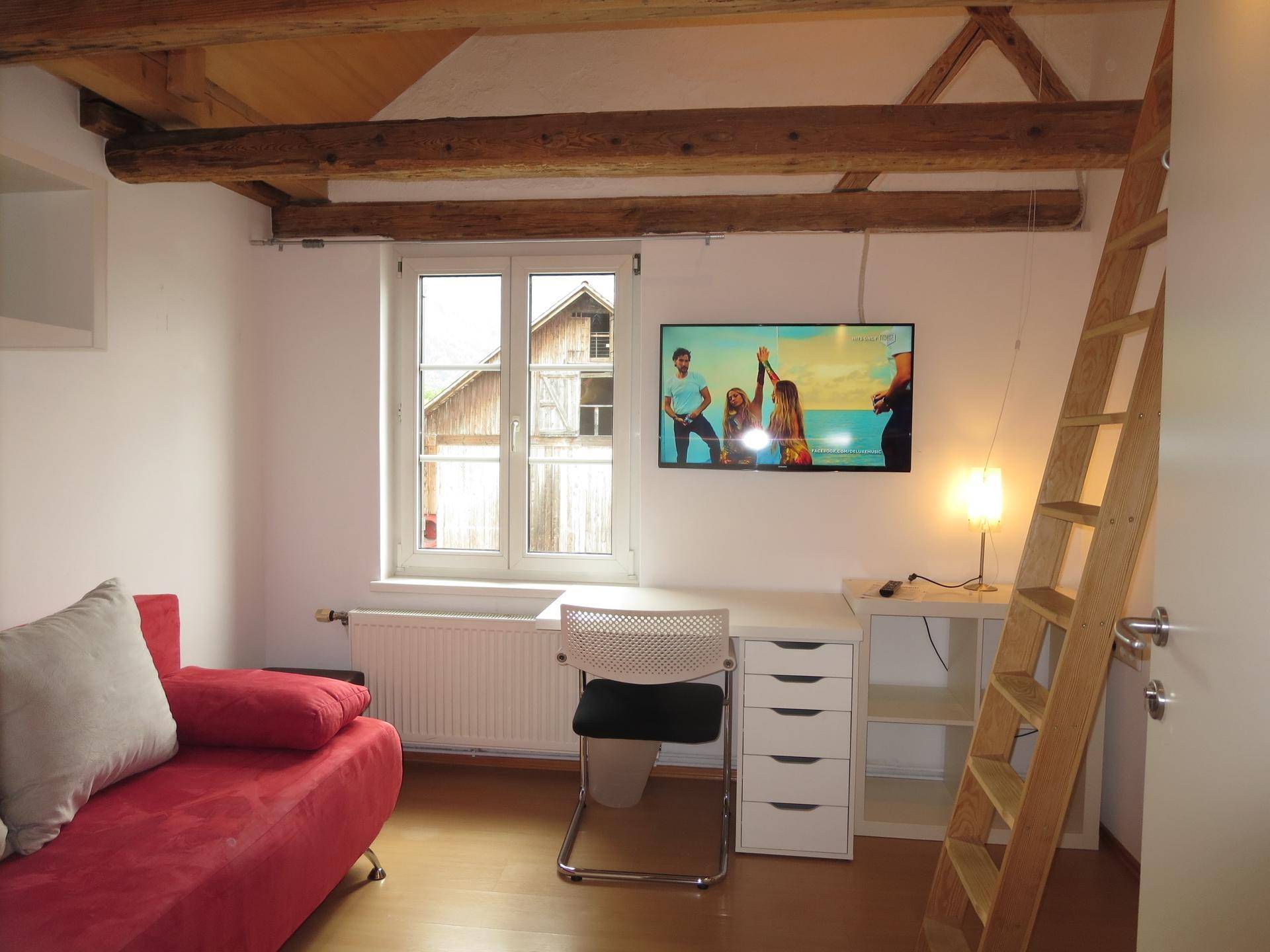 Schlafzimmer mit rotem Sofa, Schreibtisch, Fernseher und sichtbaren Holzbalken, mit Leiter zum Hochbett.