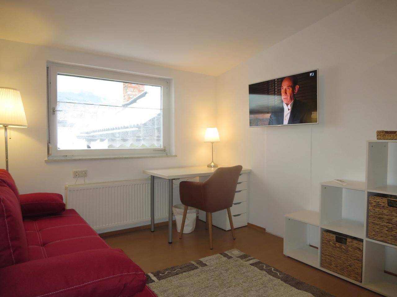 Heller Arbeitsbereich mit Schreibtisch, rotem Sofa und Blick aus dem Fenster.