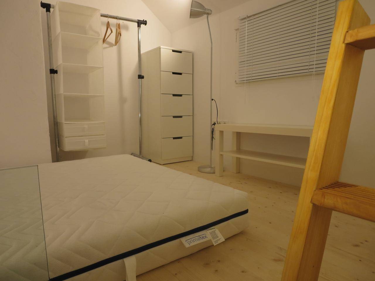 Schlafzimmer mit Matratze auf dem Boden, Kleiderstange und Leiter zum Hochbett.